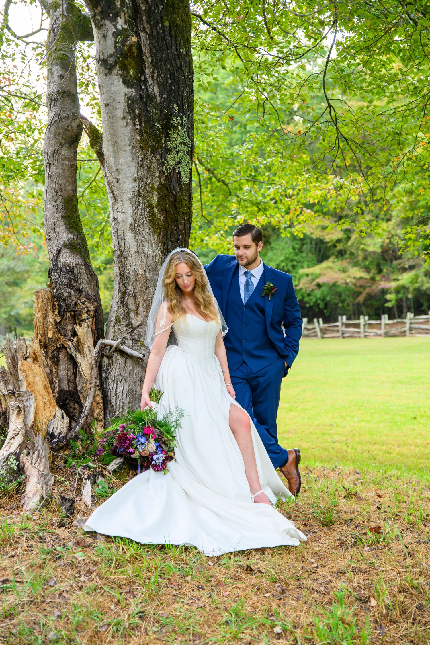 Bride in white gown holding bouquet stands with groom in navy suit under large tree on green lawn.