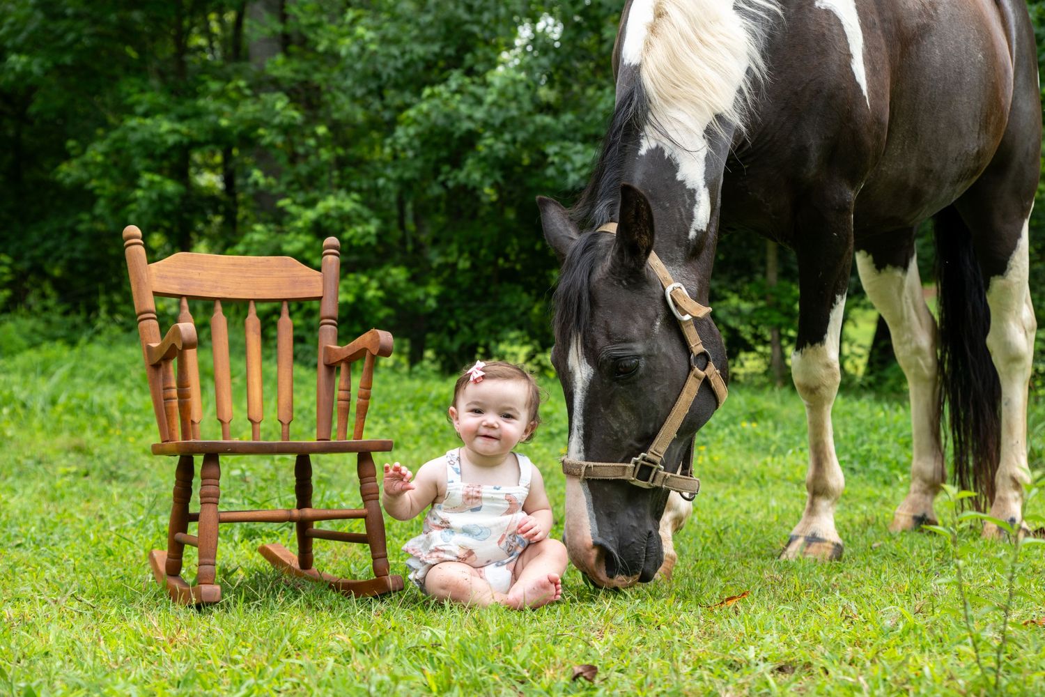 Baby girl sits on grass beside a wooden rocking chair and a grazing black and white horse outdoors.
