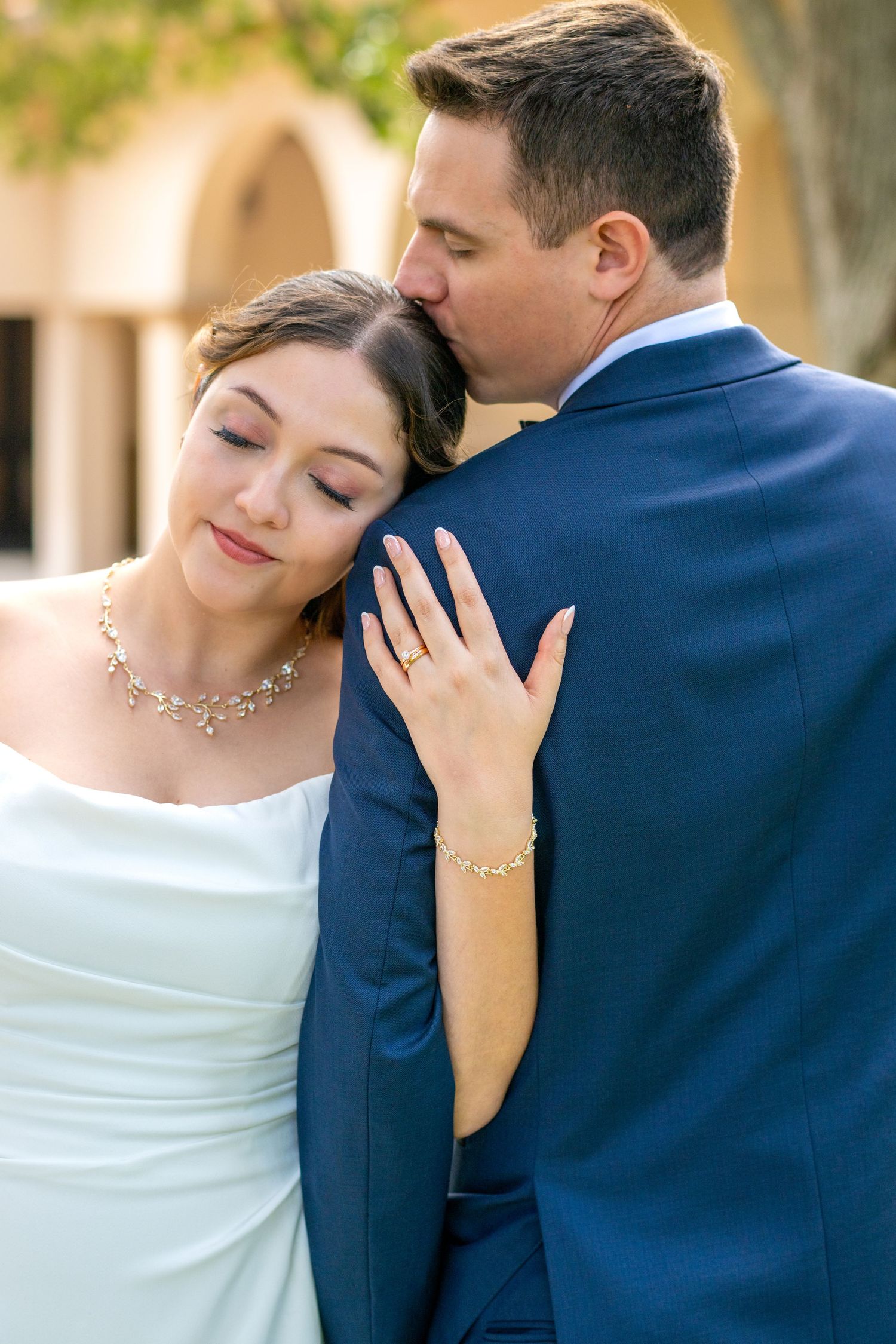 Groom in blue suit kisses bride on head as she rests hand on his chest showing wedding ring.