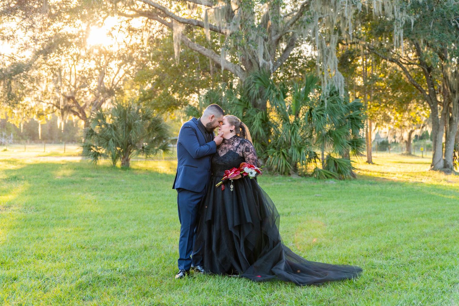 Couple shares romantic kiss in black wedding attire among moss-draped oak trees at golden hour outdoor ceremony.
