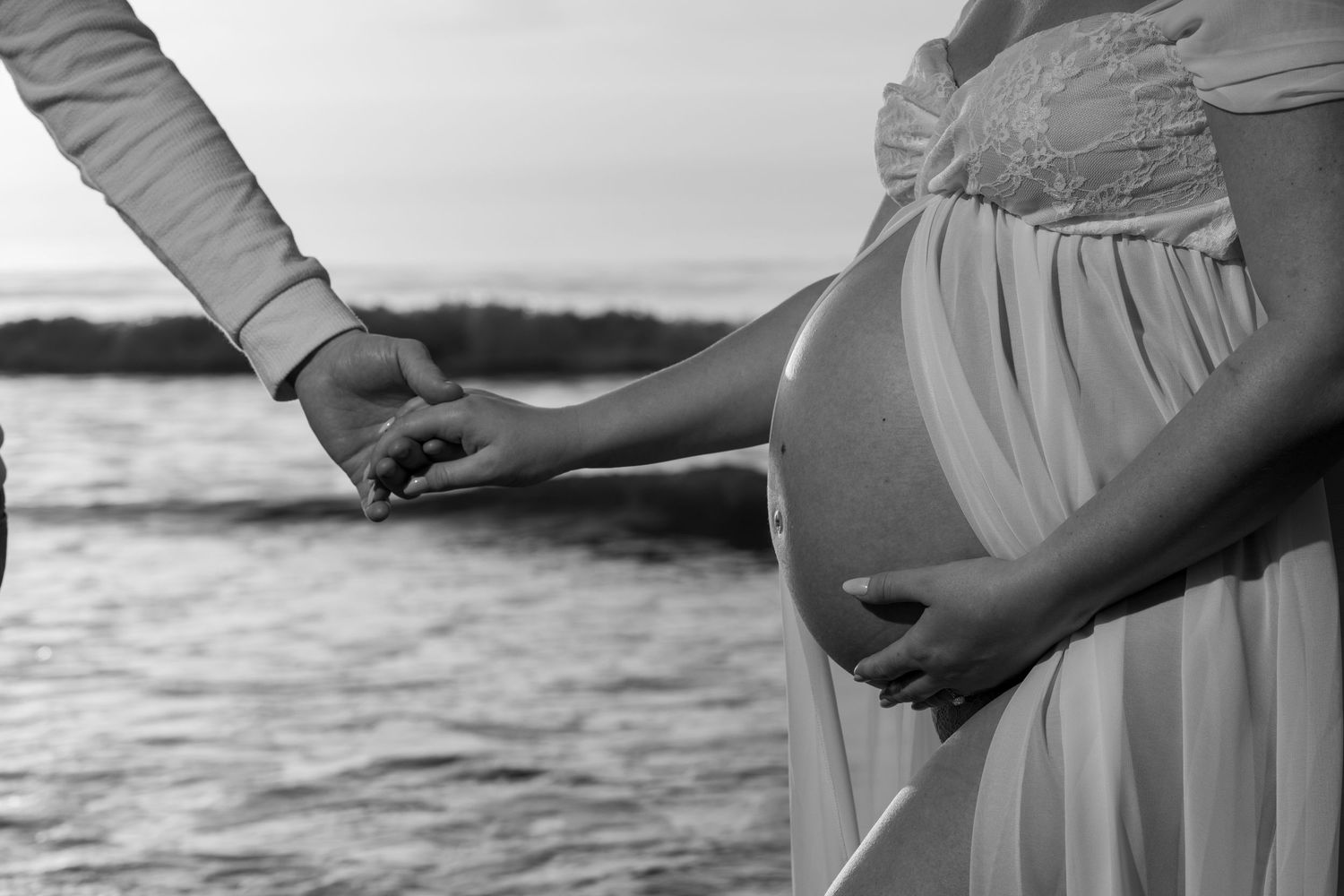 Black and white close-up of a pregnant woman holding her belly while her partner holds her hand by the ocean.