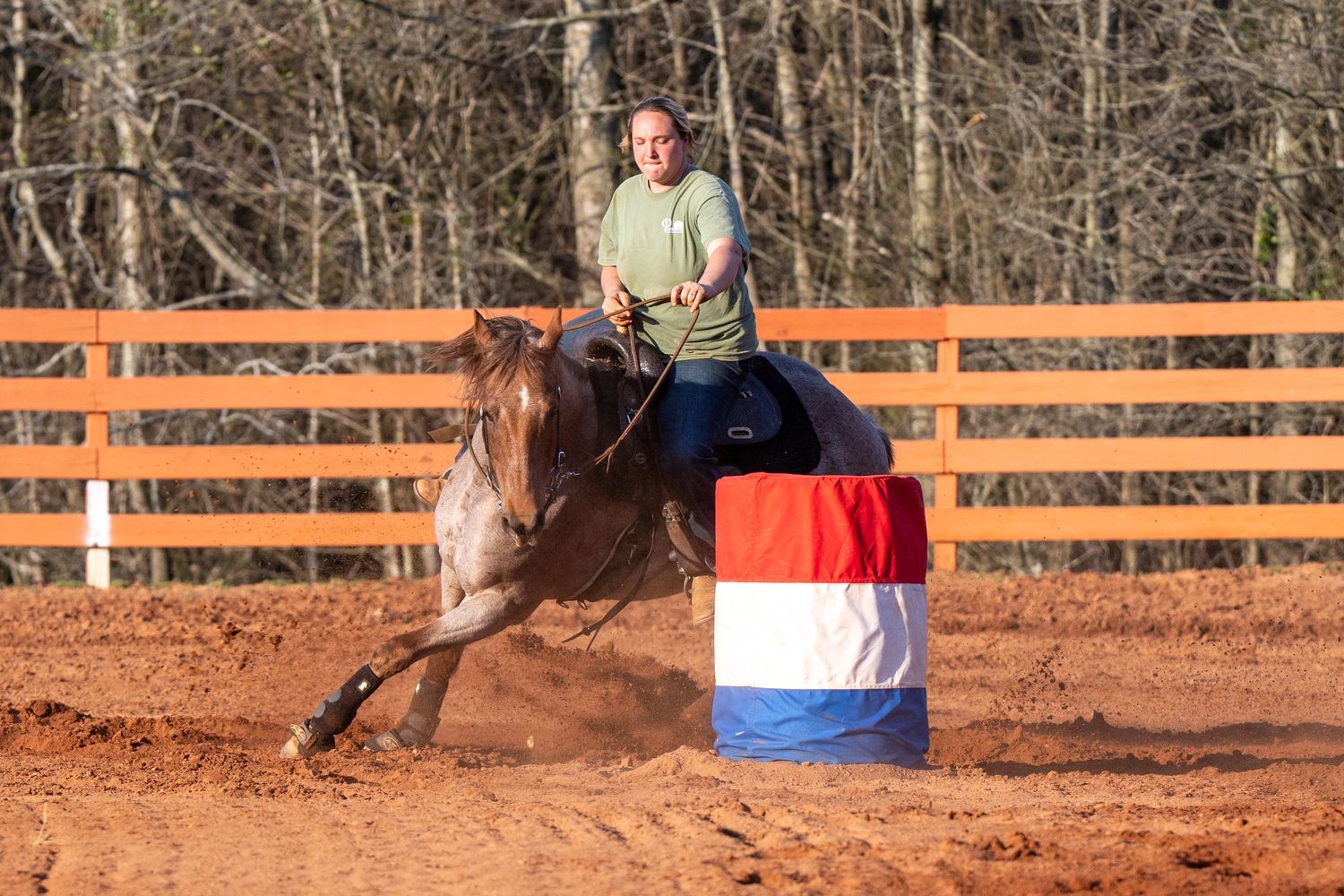 Rider on roan horse turns tight around red white blue barrel in dirt arena.