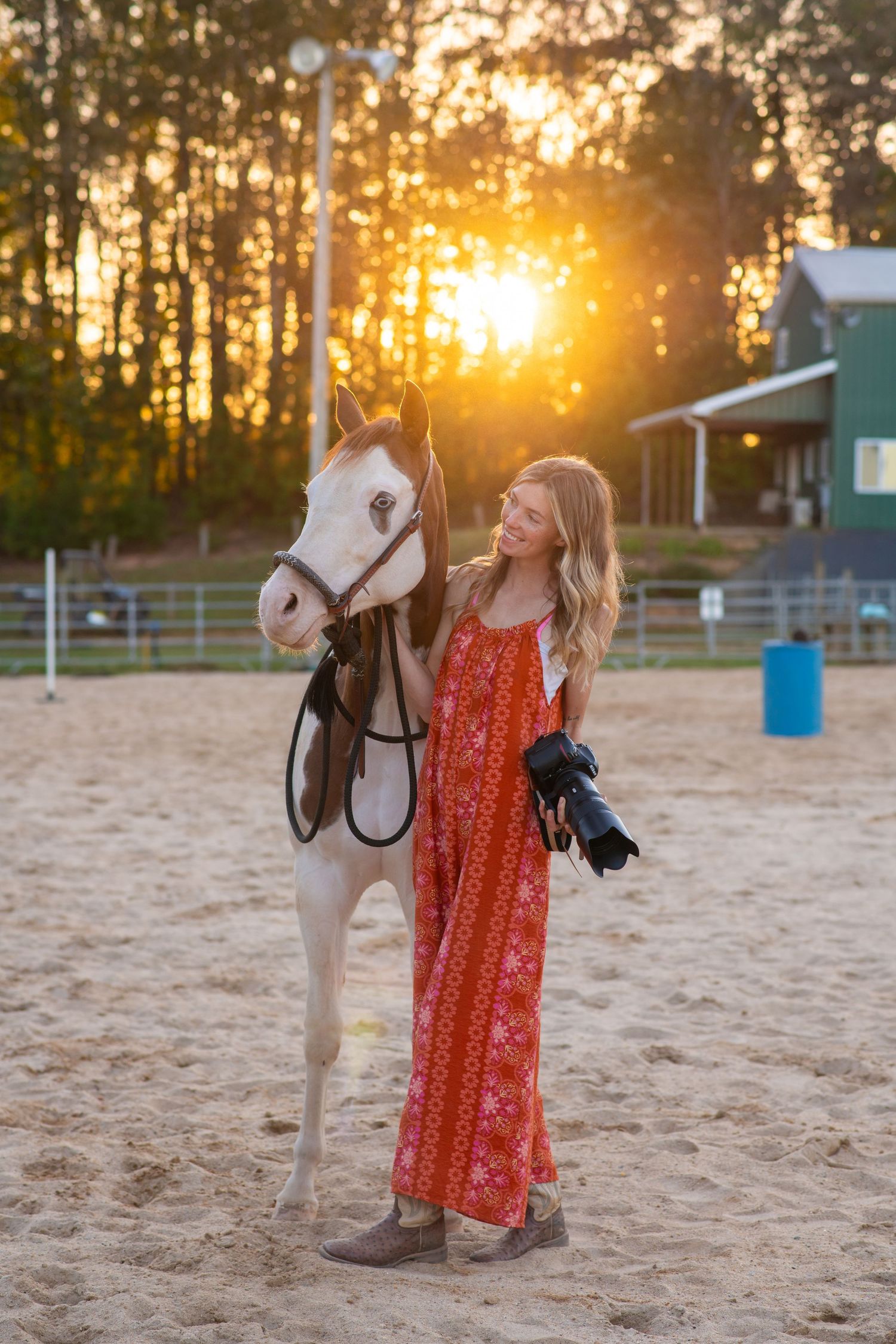 Blonde girl in red floral jumpsuit holding camera beside a pinto horse at sunset.