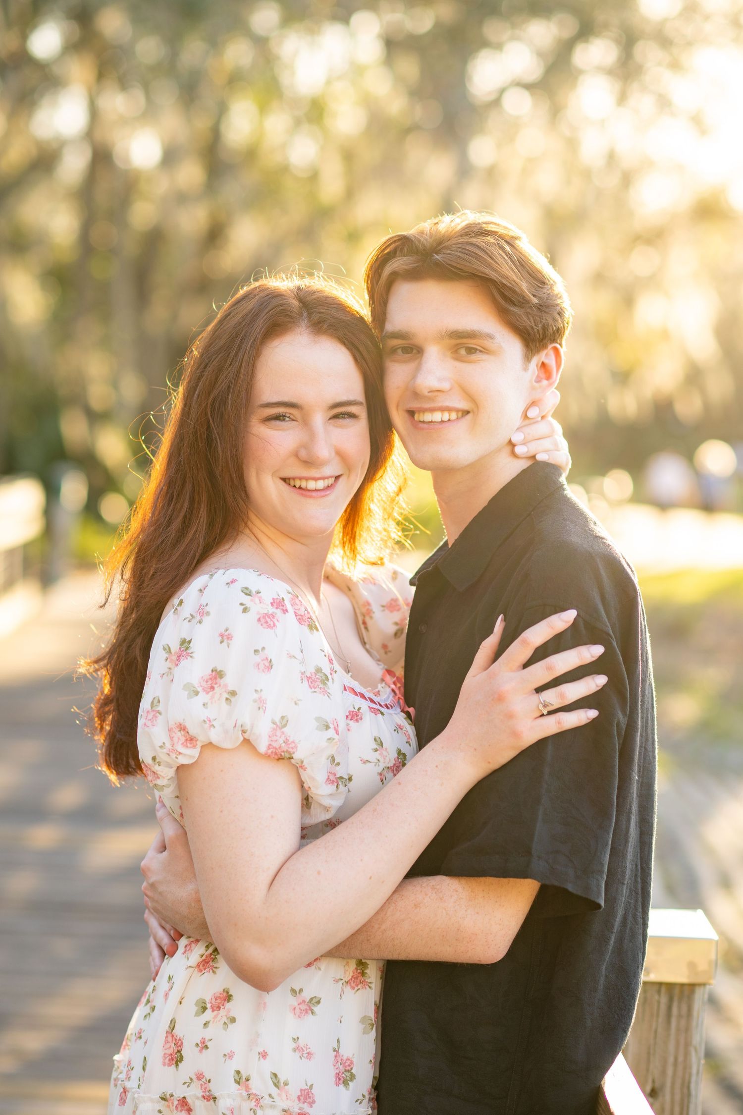 A smiling young couple embraces outdoors at golden hour, she in a floral dress, he in black, on a sunlit wooden bridge.