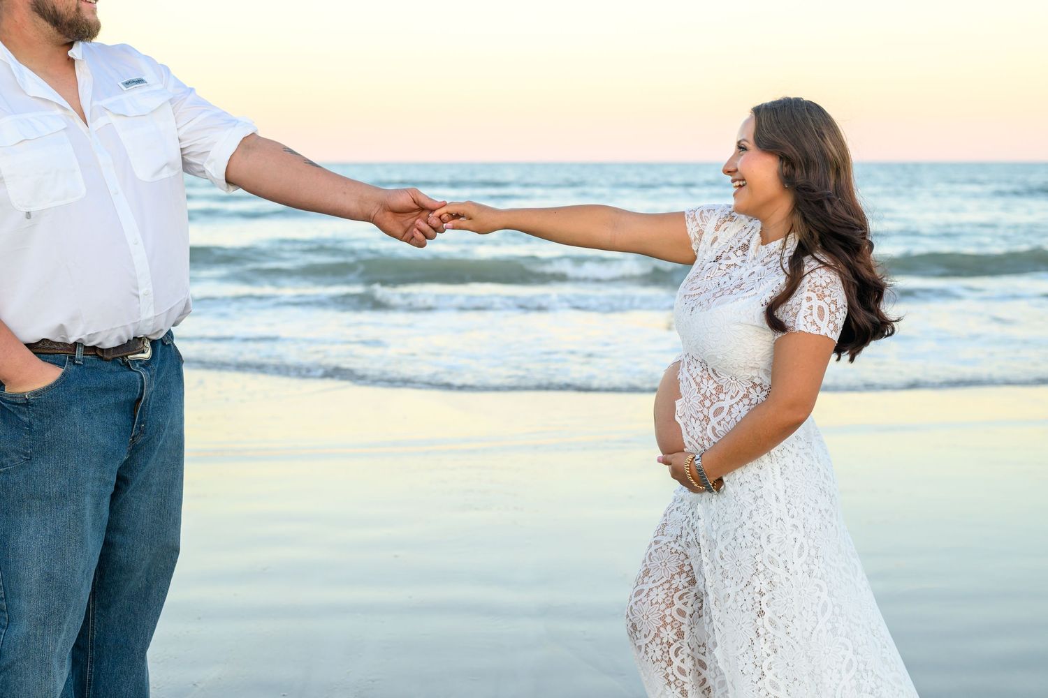 Pregnant woman in a white lace dress holds hands with a man in a white shirt and jeans on a beach at sunset.