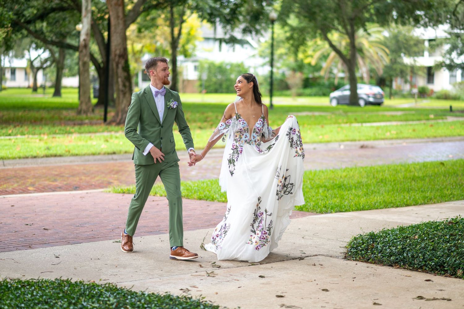 Bride in floral wedding dress and groom in green suit walking hand in hand along a sunny park path.