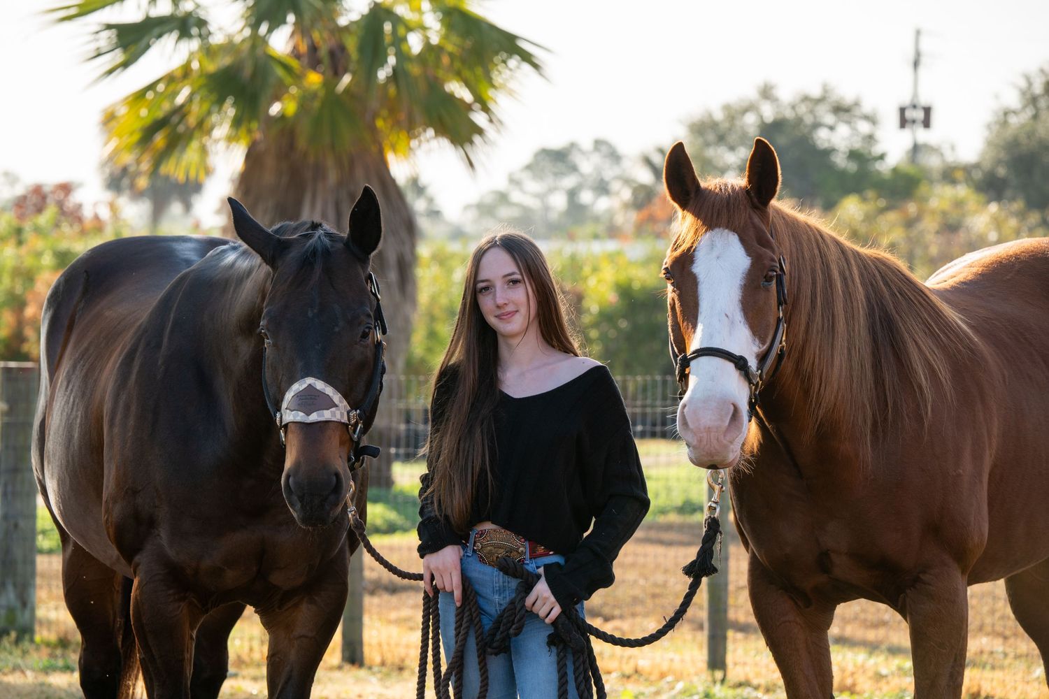Young woman with long hair stands between two brown horses on a sunny farm, holding their reins outdoors.
