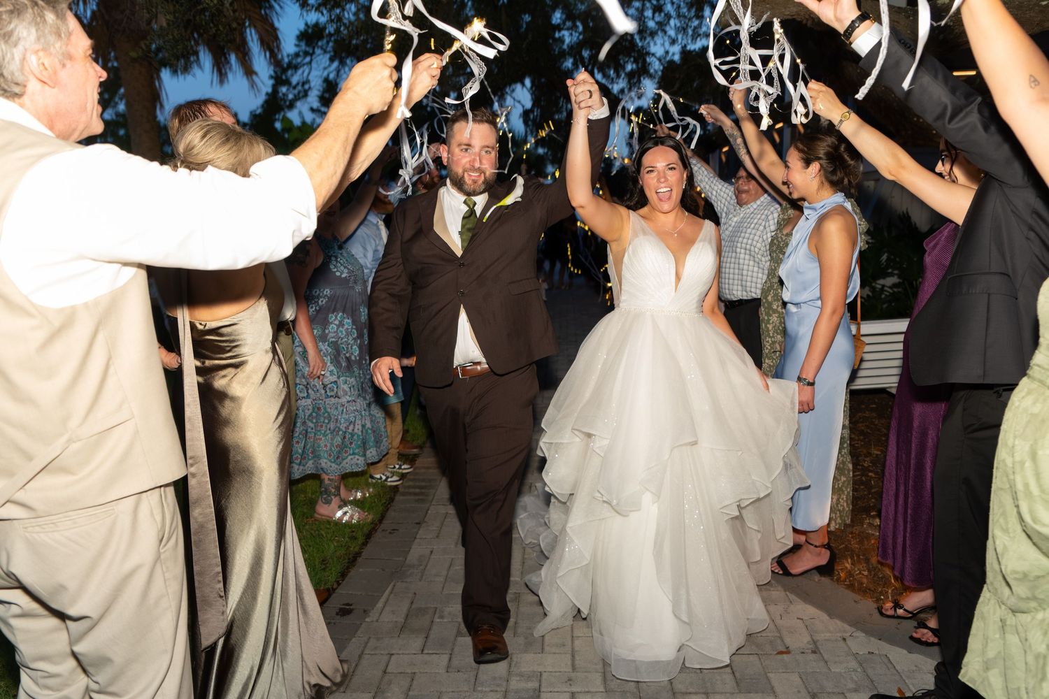 Bride and groom exit wedding reception through a celebratory ribbon wand sendoff surrounded by cheering guests at night.