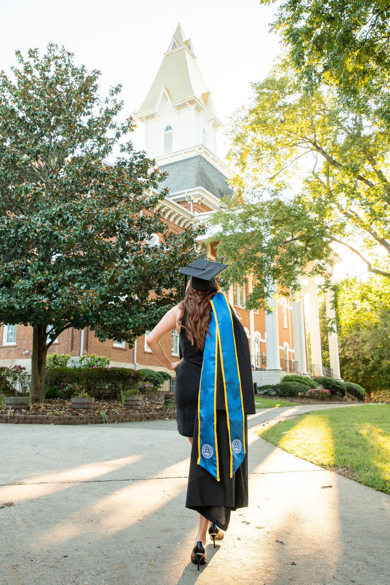 Graduate in black gown and blue stole walks toward historic brick building on sunny campus path.