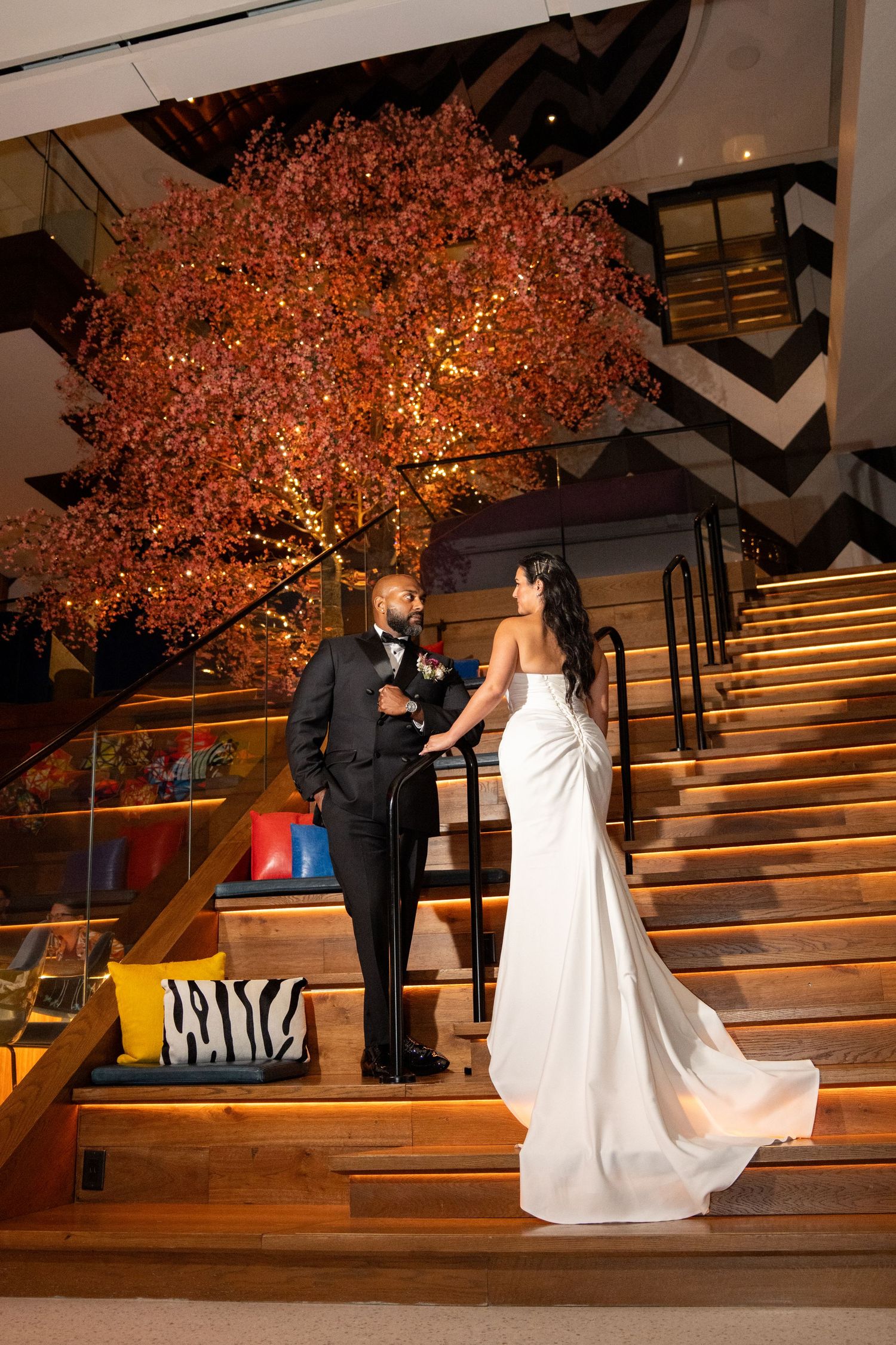 Elegant bride and groom hold hands on grand staircase beside illuminated autumn tree at wedding venue.