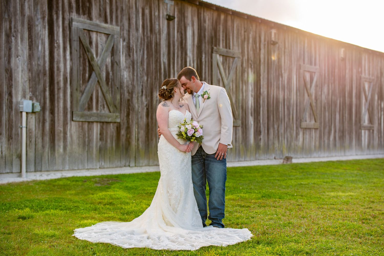 Bride and groom share intimate moment in front of rustic wooden barn in warm golden sunlight.