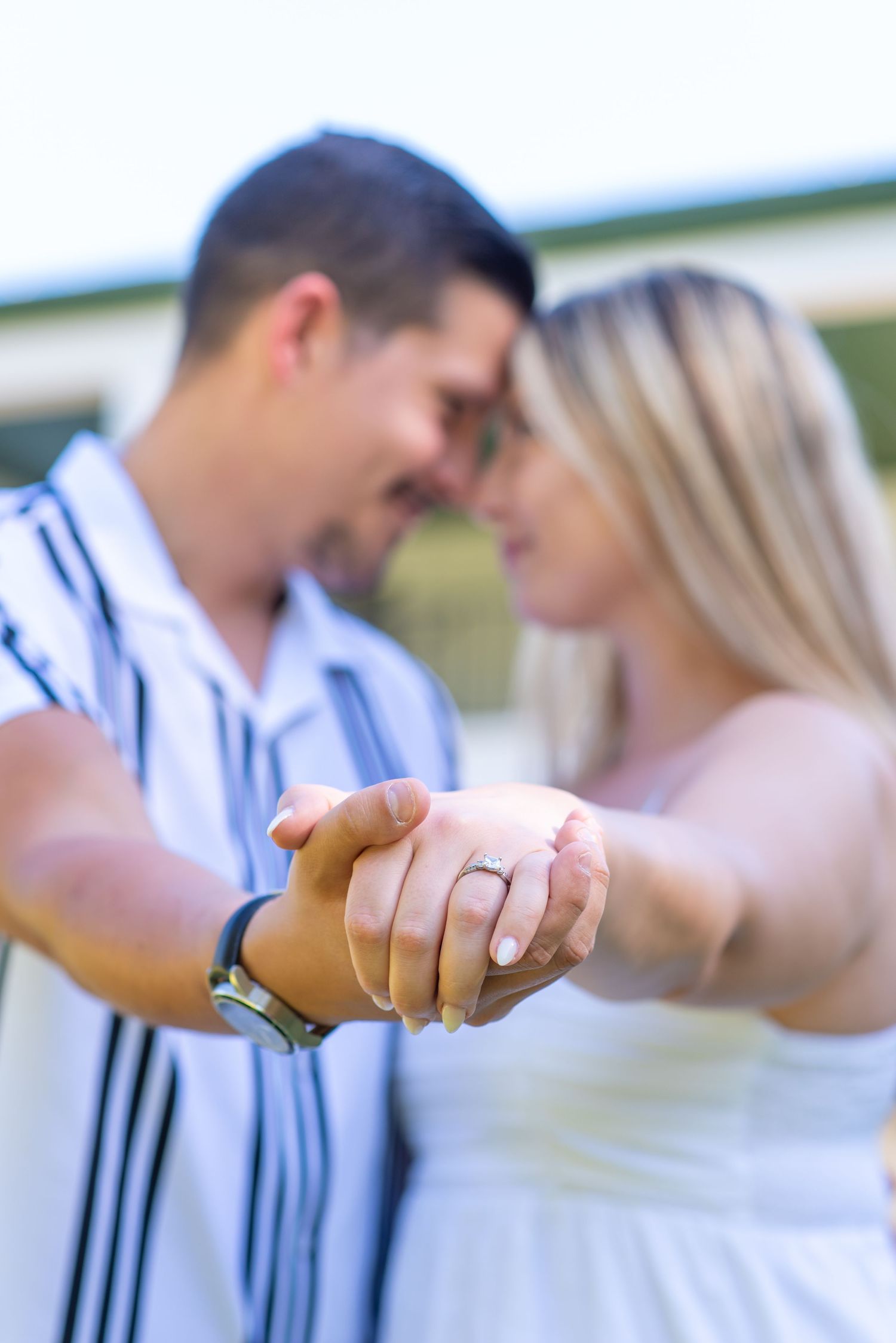 Engaged couple holding hands toward camera, woman displaying diamond engagement ring, foreheads touching.
