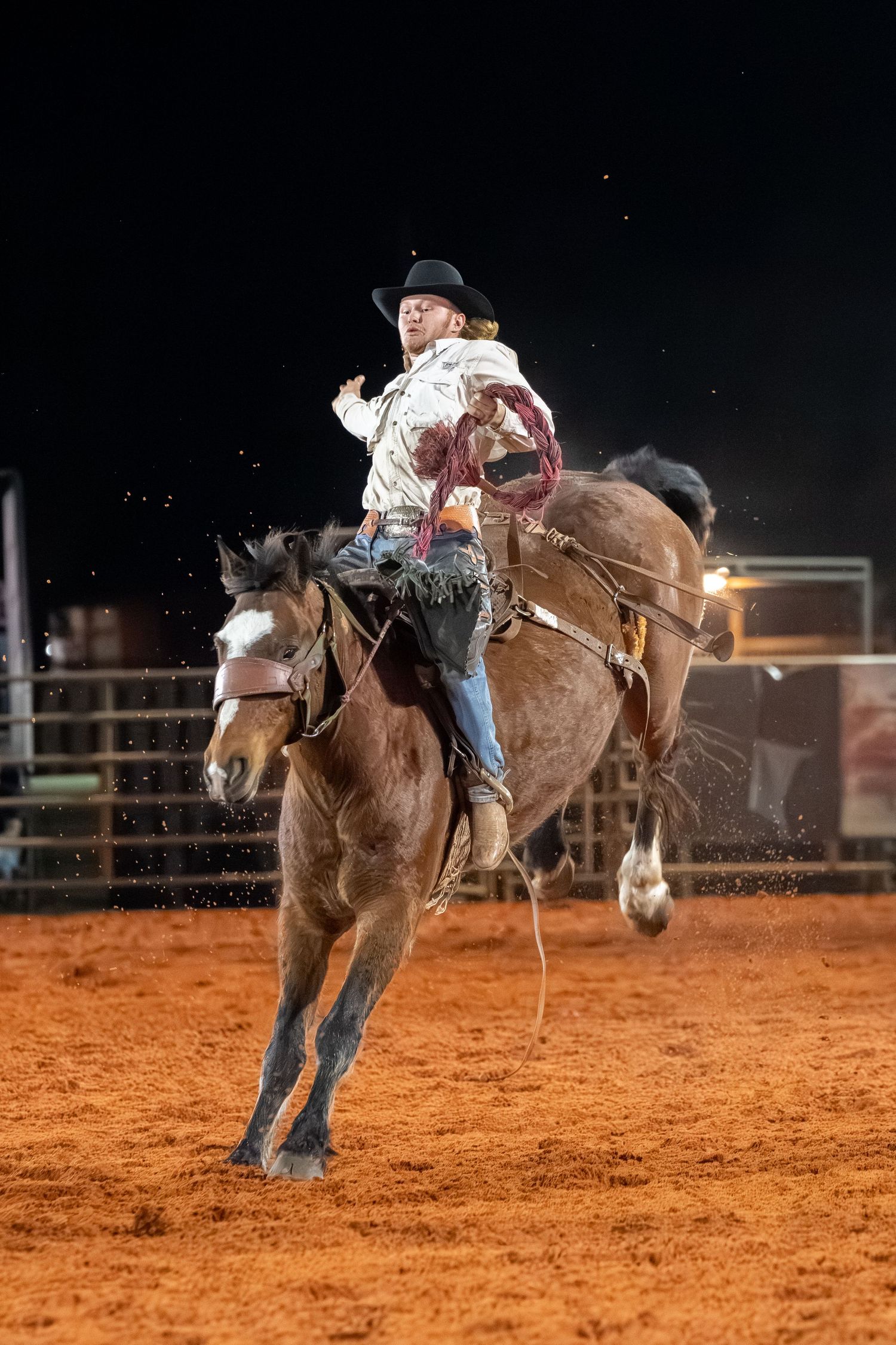 Cowboy in white shirt rides a bucking brown horse during a nighttime rodeo bronc riding competition.