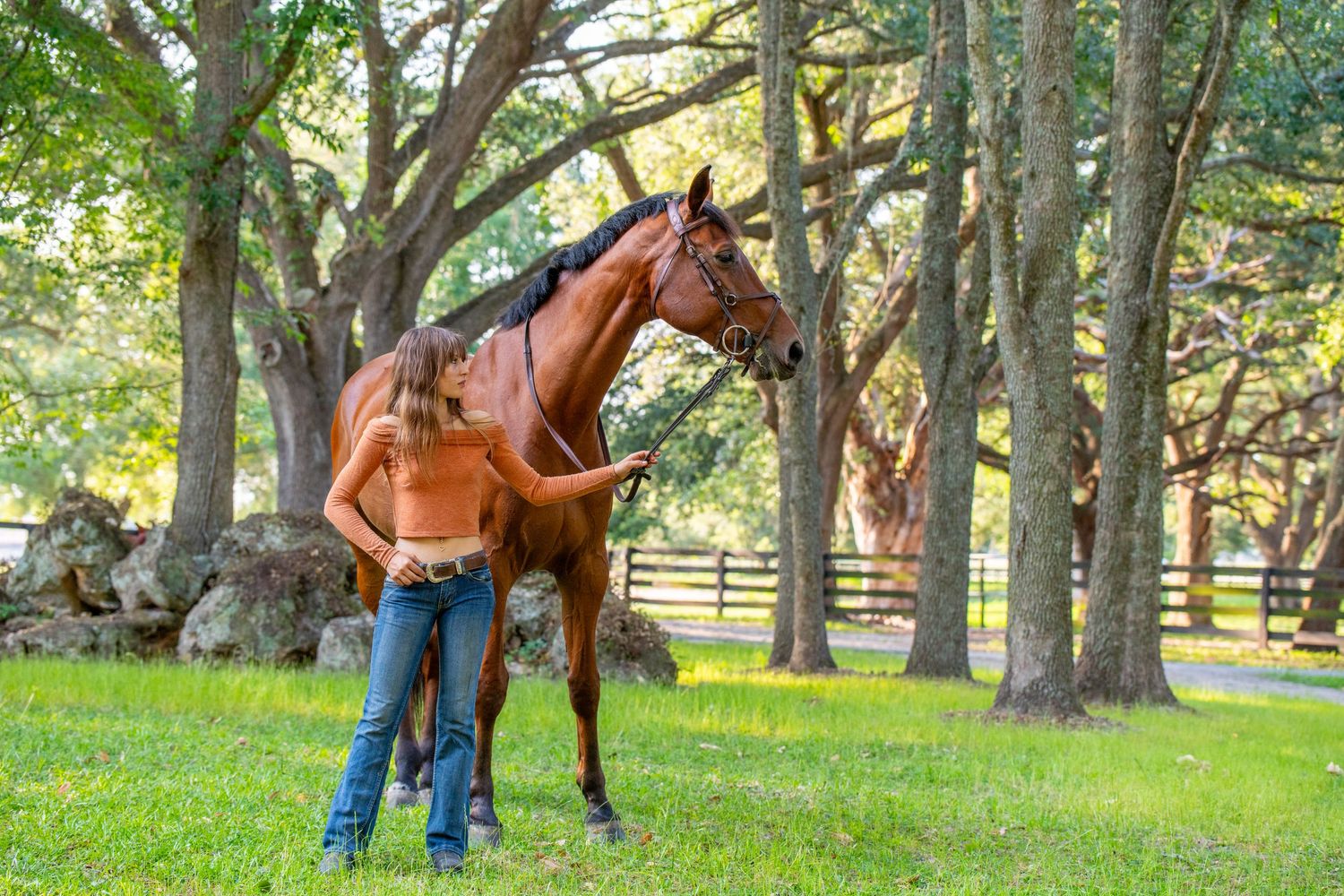 Woman in crop top standing with a chestnut horse in a lush green tree-lined pasture.