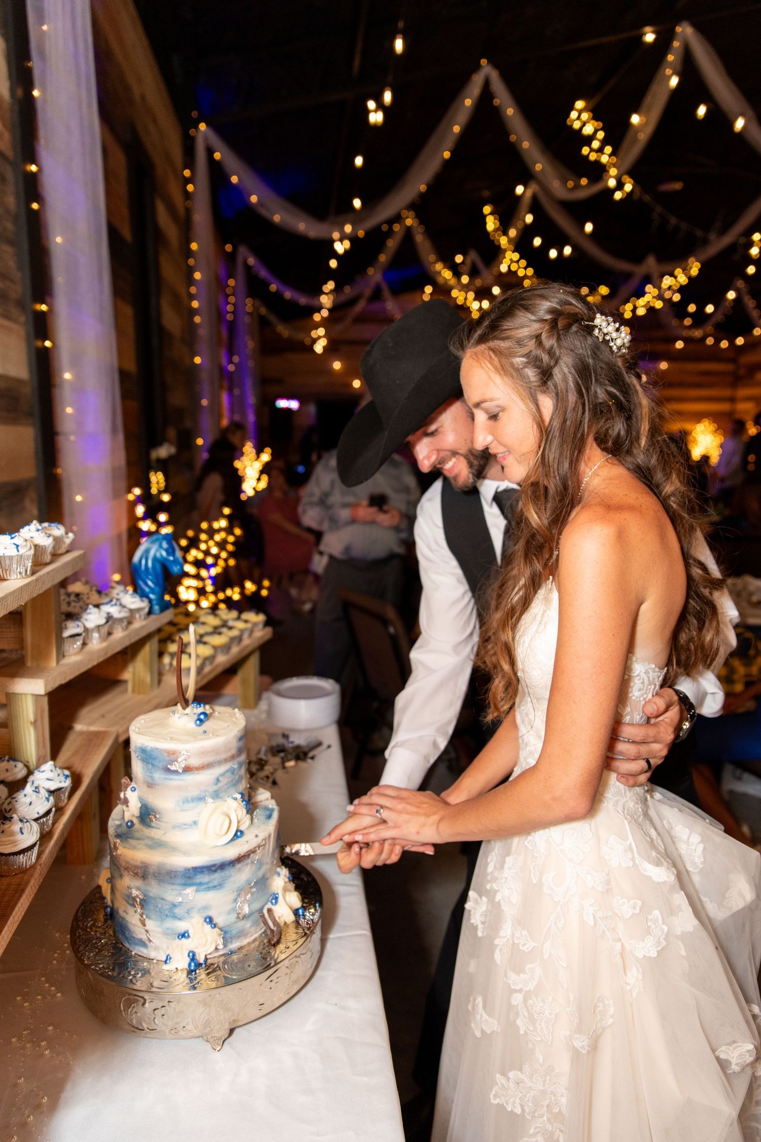 Bride and groom cut a blue and white tiered wedding cake under warm string lights in a rustic barn.