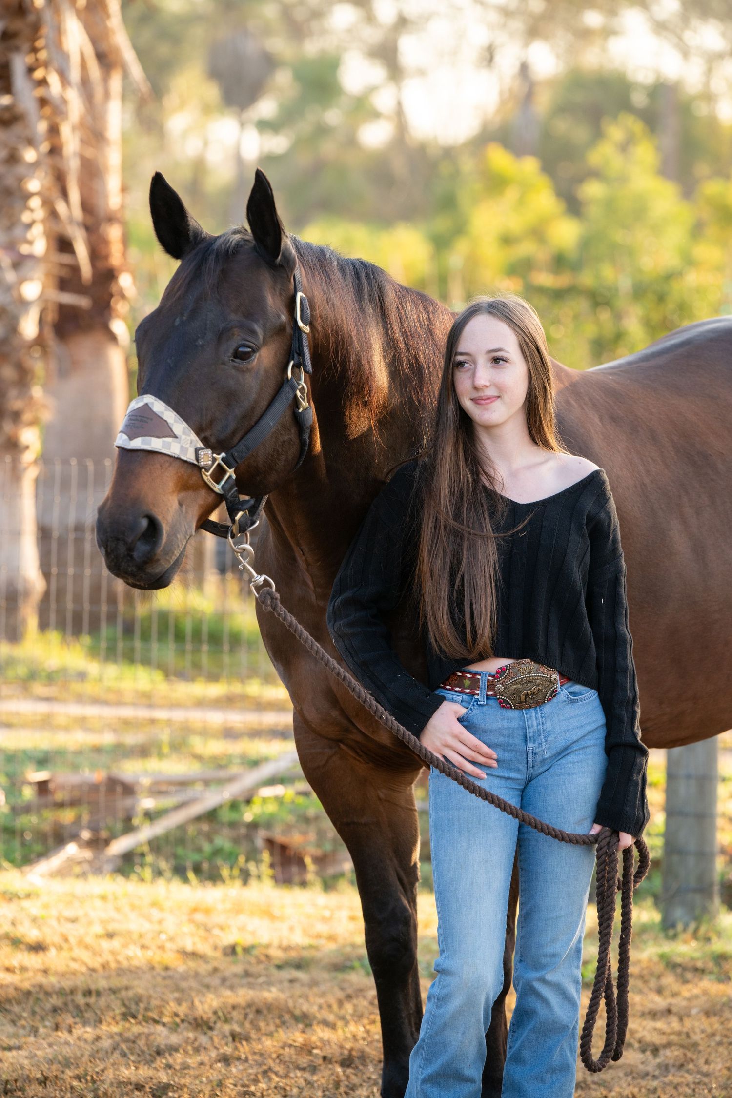 Young woman with long hair in jeans and black top stands beside a dark brown horse on a sunny farm.