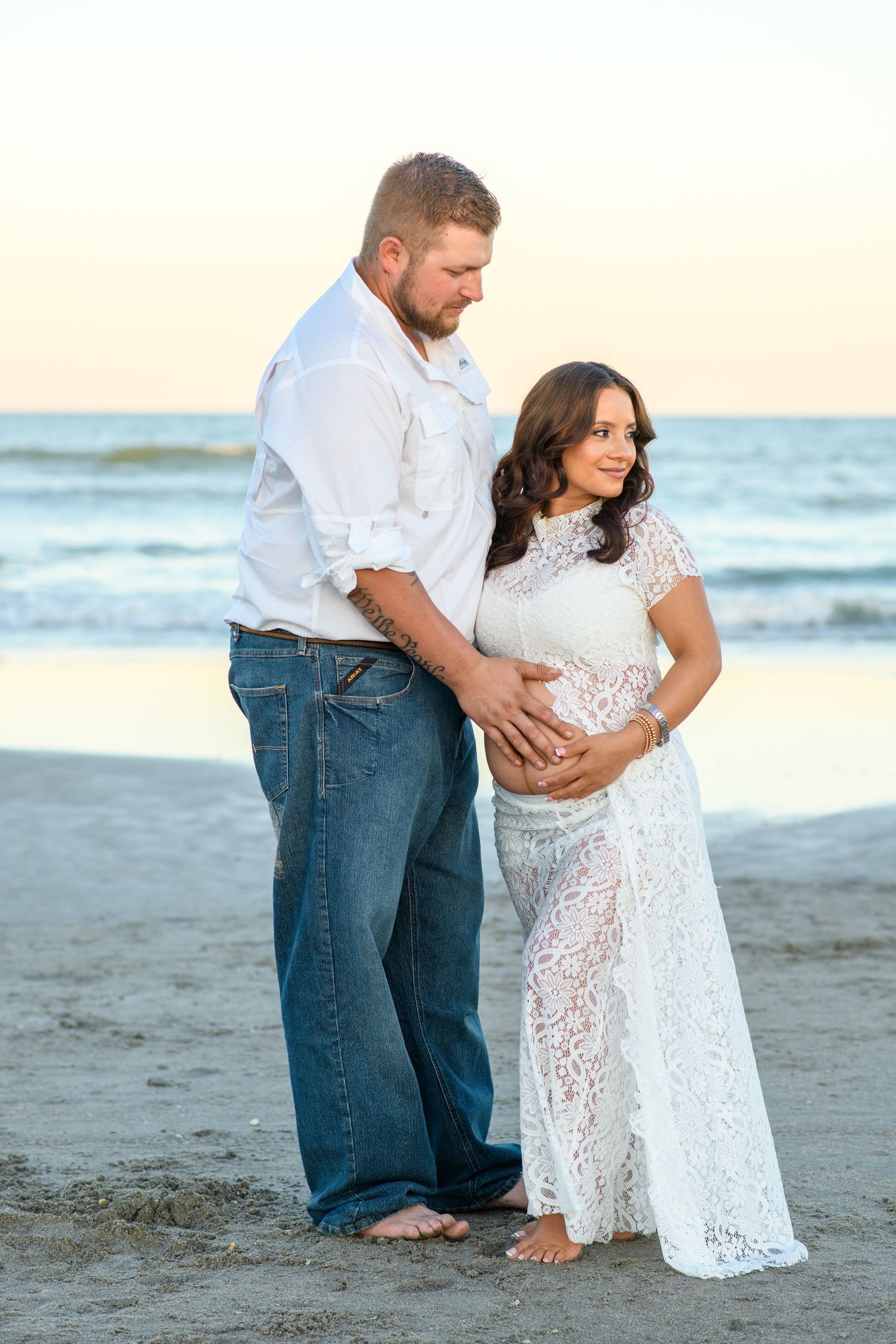 Expecting couple in white clothing embrace on a sandy beach at sunset, the man cradling the woman's baby bump in a lace dress.