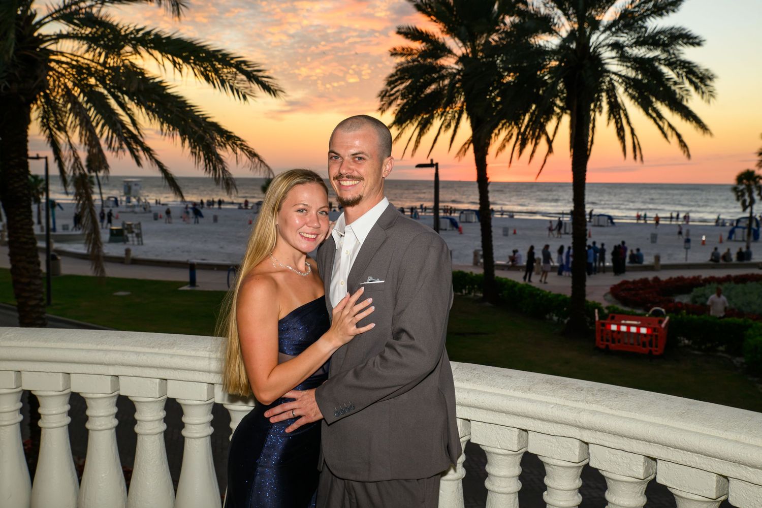 Smiling couple in formal attire embrace on a white balustrade terrace with palm trees, a beach, and a colorful sunset sky behind them.