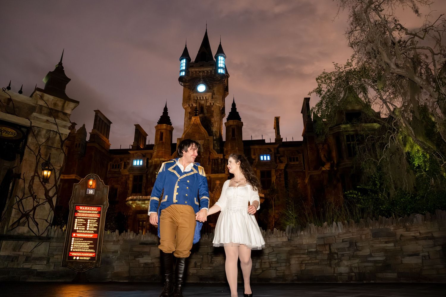 Couple in Beauty and the Beast costumes posing in front of a castle at night.