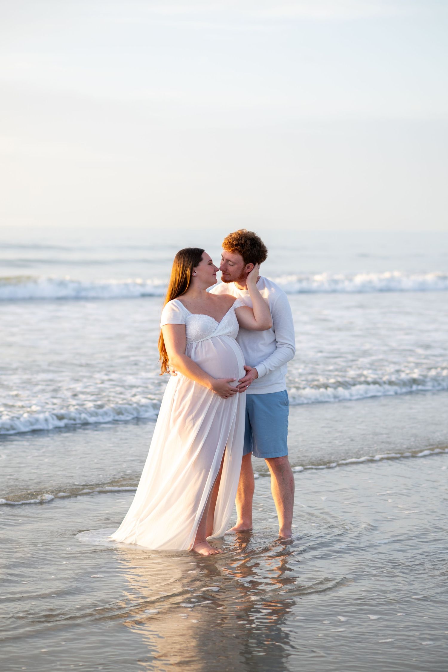 Pregnant couple embracing on beach at sunset, woman in white flowing gown, man in white shirt and blue shorts, ocean waves behind.