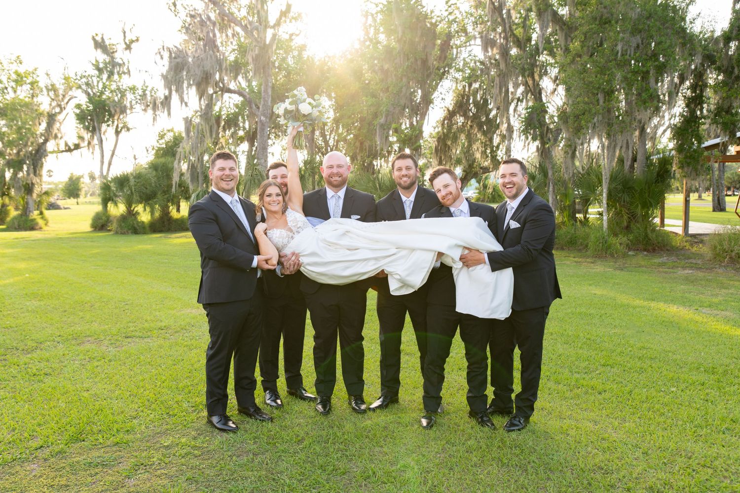 Groomsmen in black suits playfully lift bride in white wedding dress outdoors under Spanish moss trees.