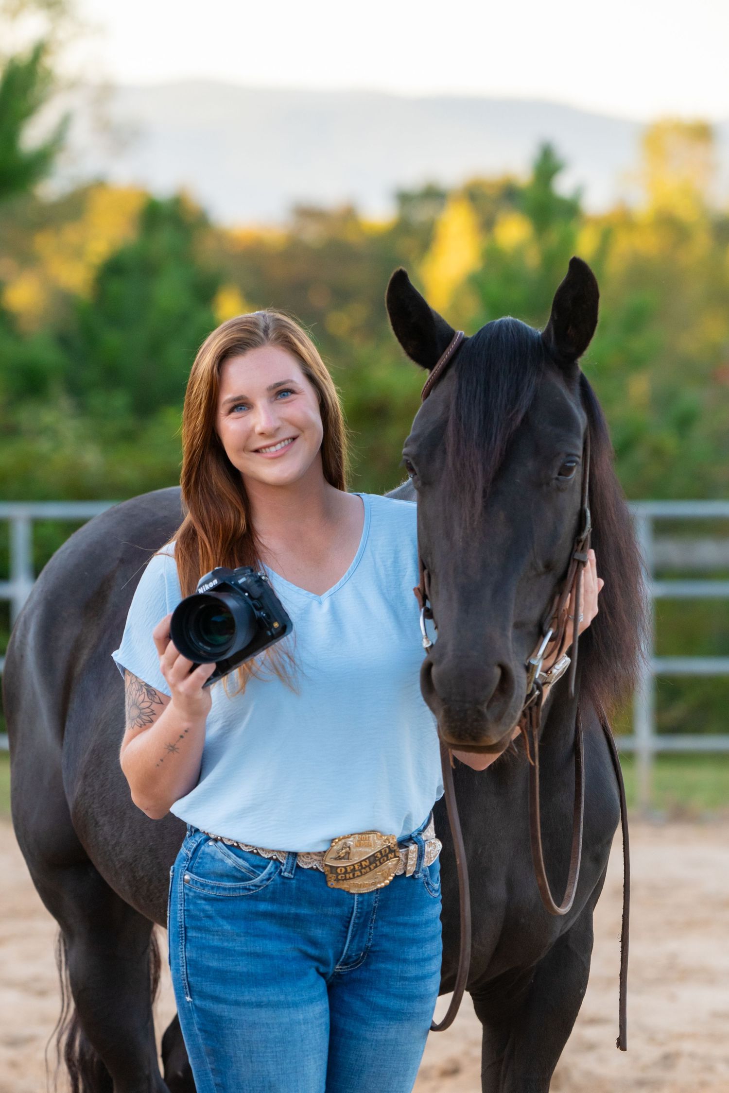 Young woman photographer holding DSLR camera stands beside dark horse in outdoor arena with mountain backdrop.