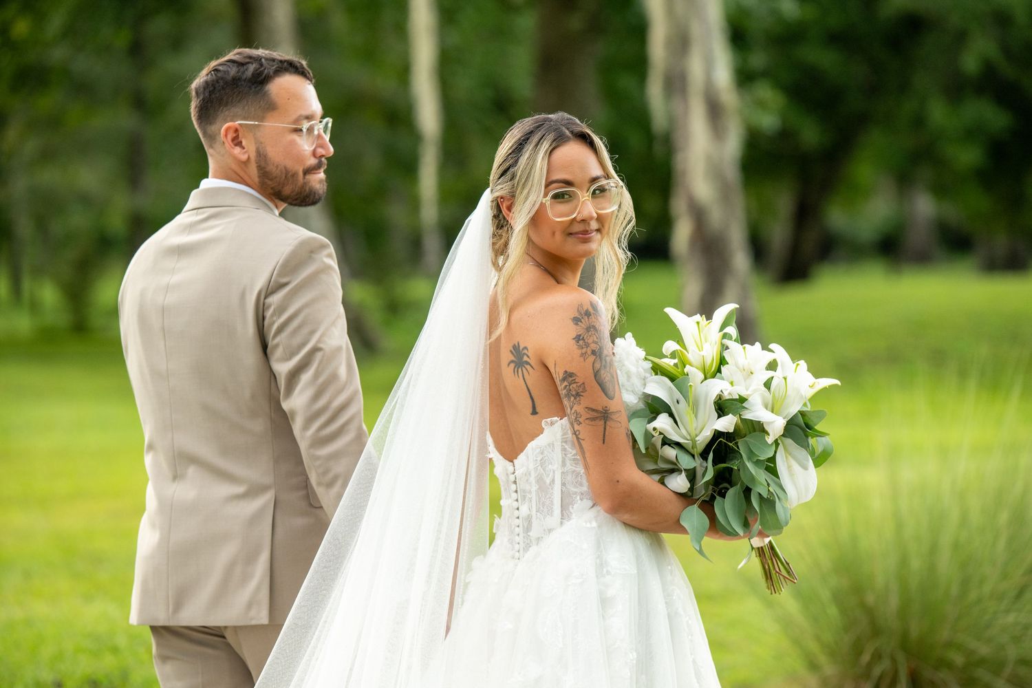Bride in white lace gown with veil holds white floral bouquet beside groom in tan suit outdoors.
