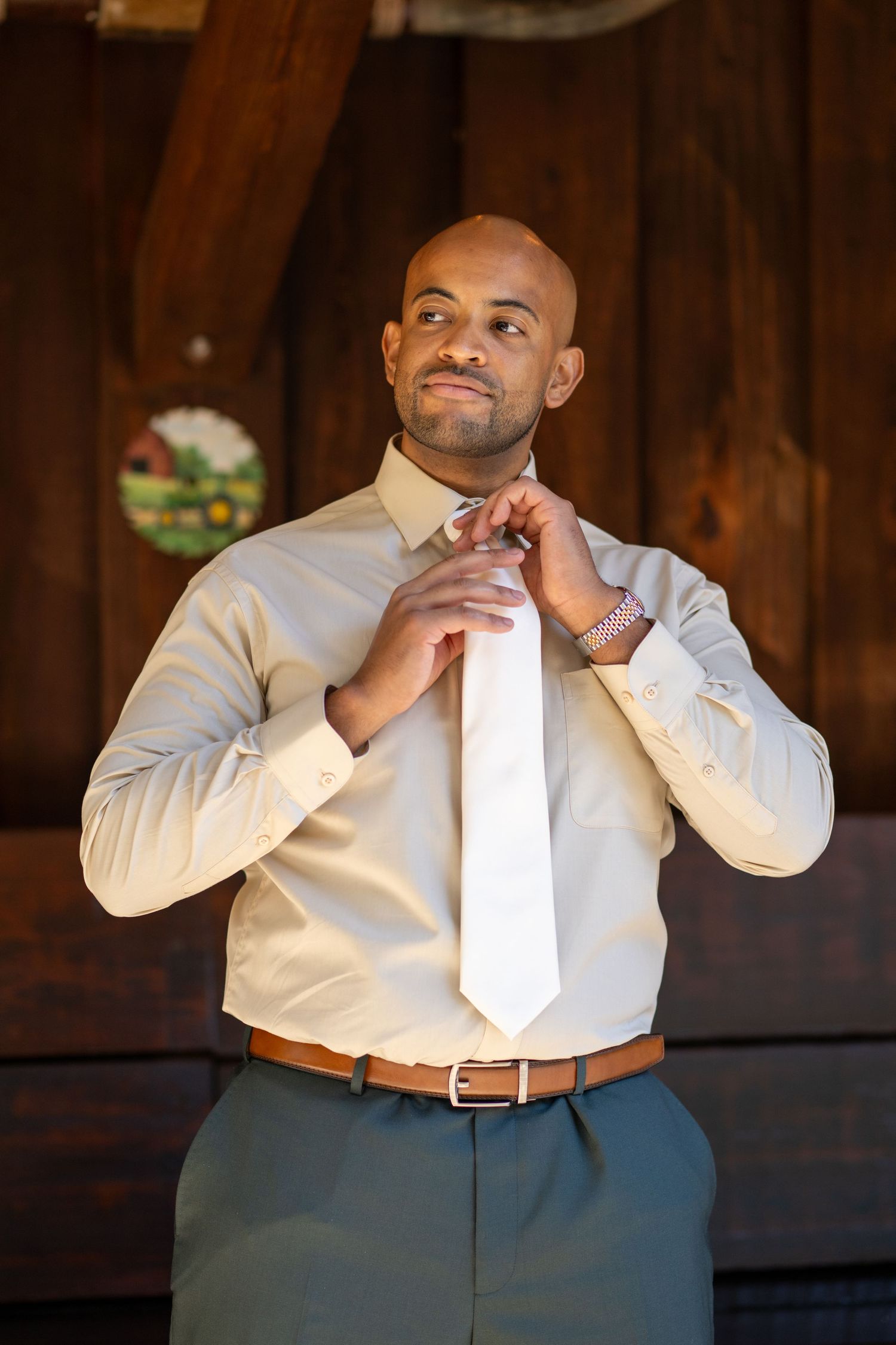Bald groom adjusting white tie over cream dress shirt with brown belt in rustic barn setting.