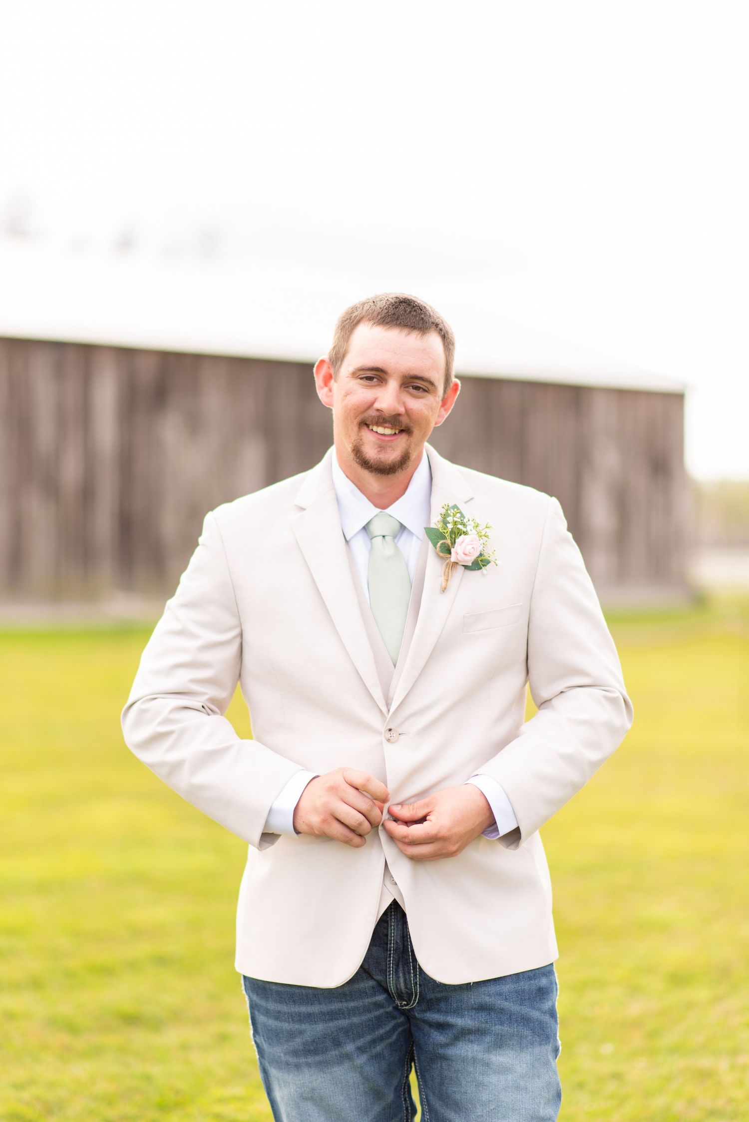 Smiling groom in beige blazer, mint tie, and jeans with boutonniere stands outdoors near a barn.