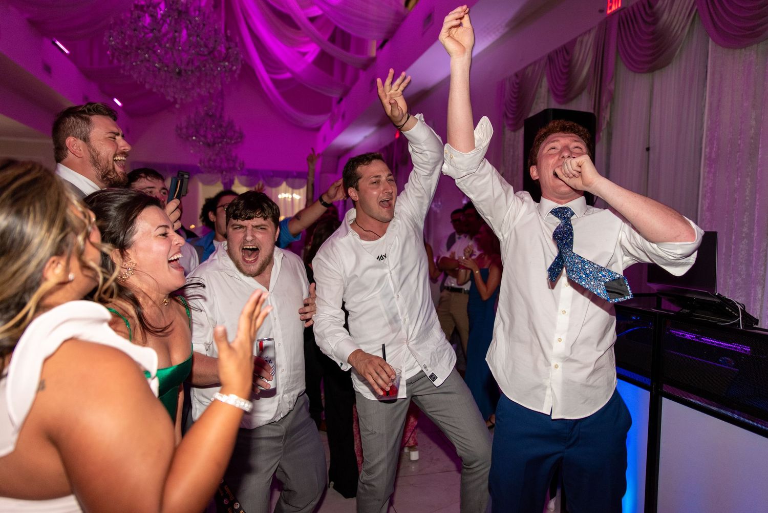 Energetic wedding guests dancing and celebrating under purple lights on the reception dance floor.