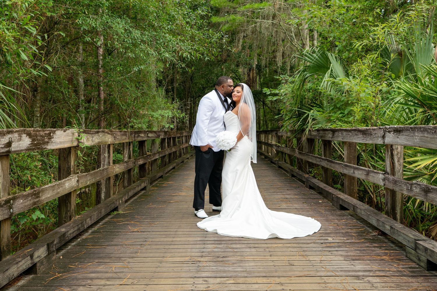 Bride and groom embracing on a wooden bridge surrounded by lush green trees in an outdoor wedding portrait.