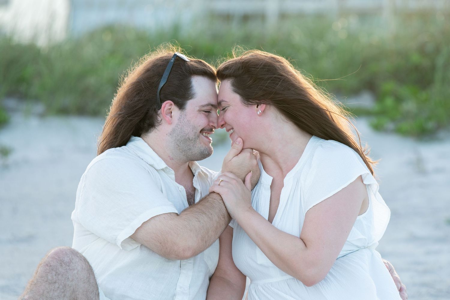 Romantic couple in white outfits touching foreheads and holding hands on a sandy beach.
