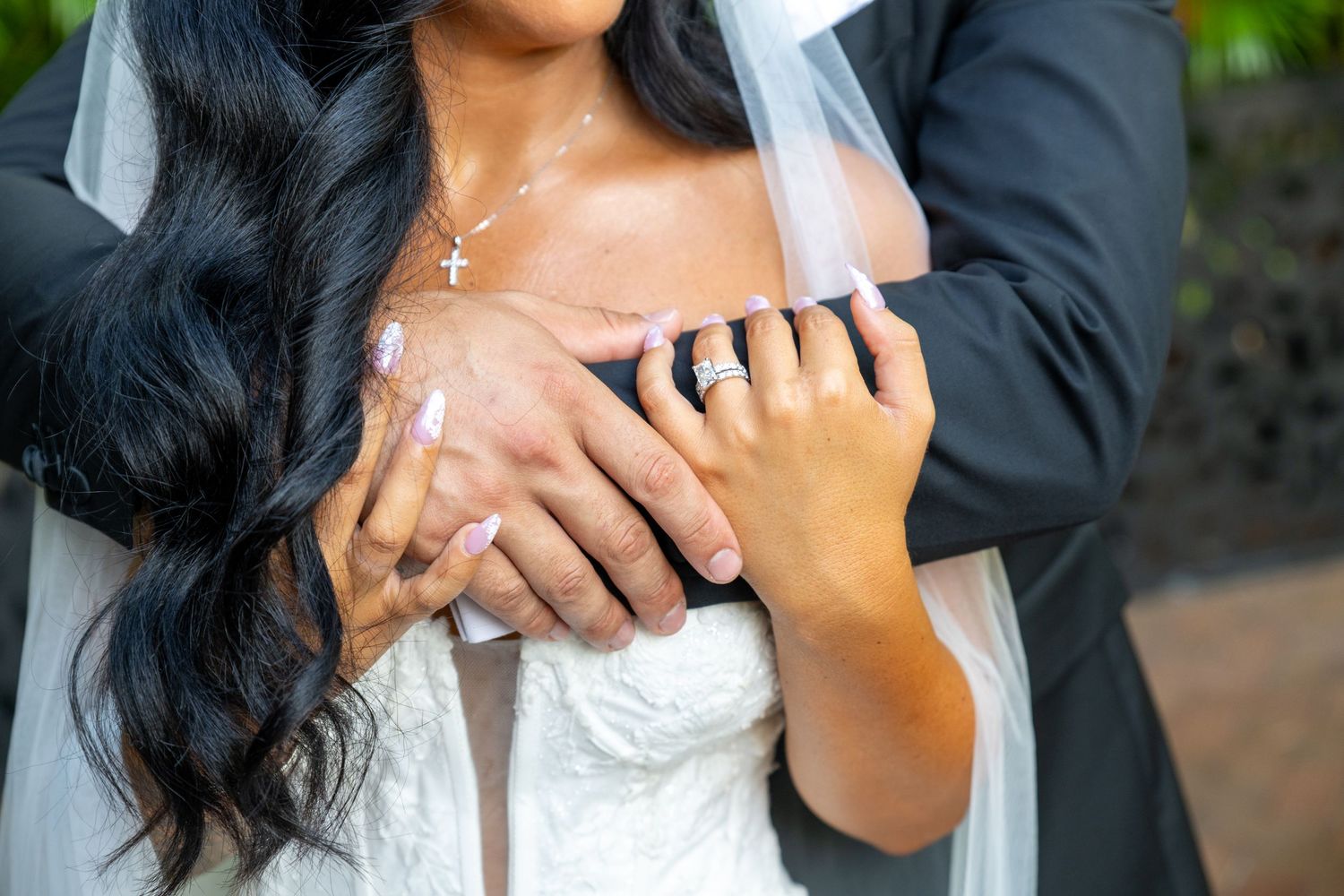 Close-up of bride and groom's hands showing wedding rings, bride wearing white lace gown and cross necklace.
