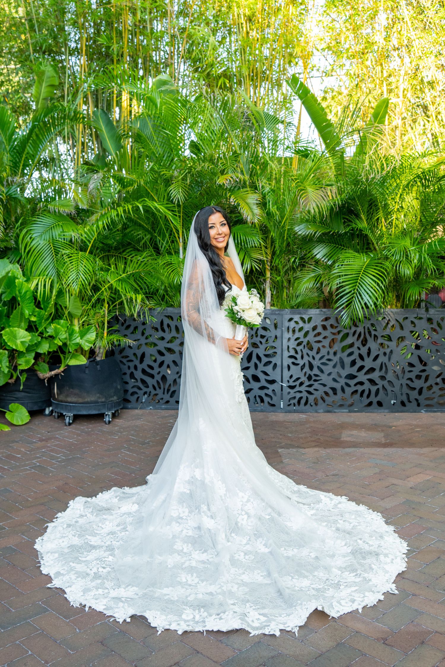 Bride in white lace gown with cathedral veil holding bouquet, posing in tropical garden setting.