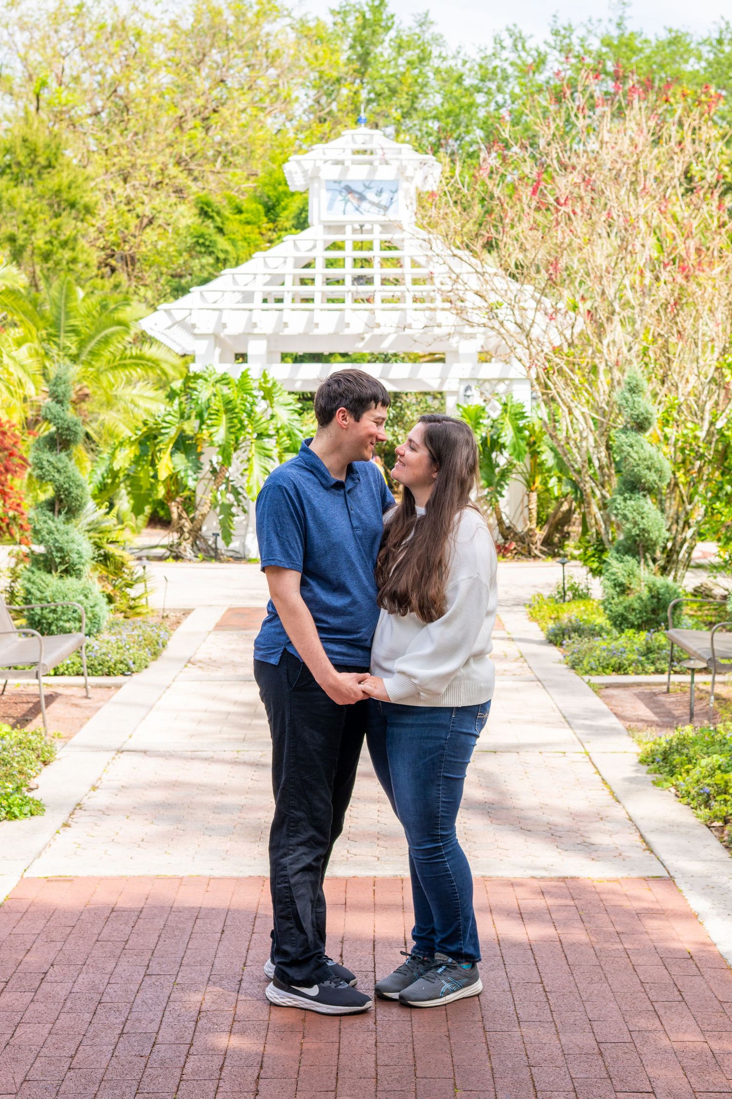 Couple holding hands and smiling at each other on a brick path in a lush garden with a white gazebo.