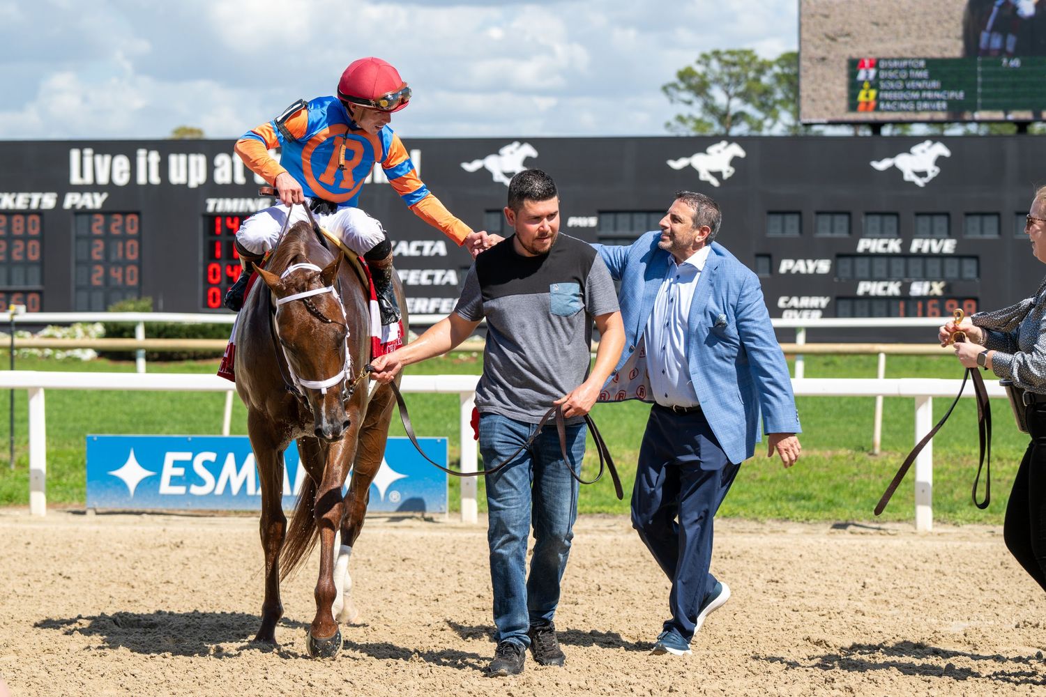 Winning jockey on chestnut horse is greeted by two men near a scoreboard after a horse race victory.