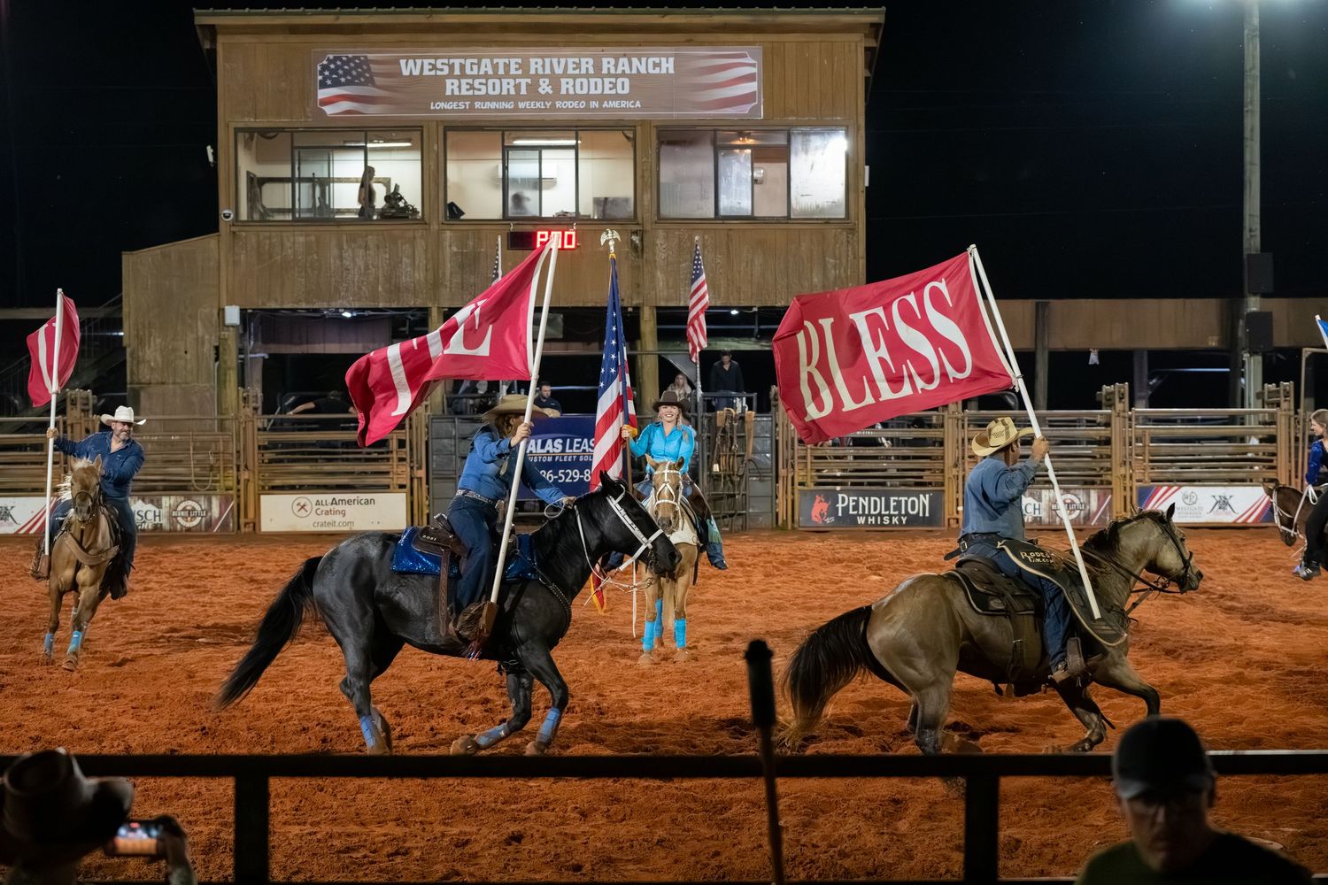 Rodeo performers on horseback carrying flags at night in illuminated indoor rodeo arena.
