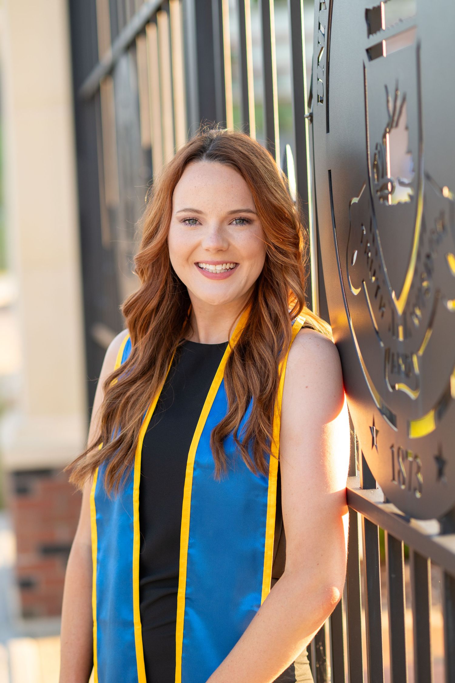 Smiling young woman in blue and gold graduation stole leaning against ornate university gate for graduation photo.