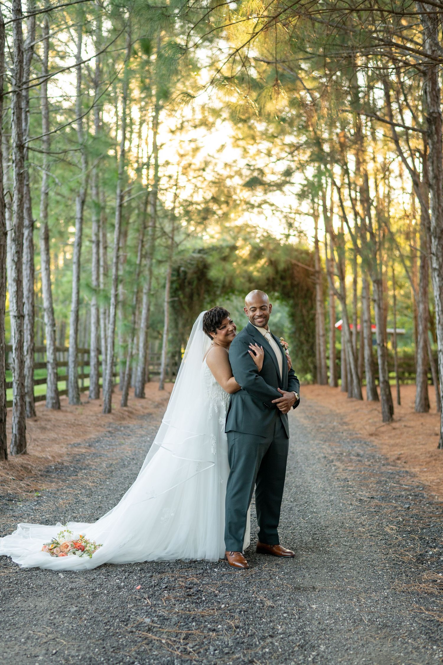 Bride and groom pose on a tree-lined gravel path at golden hour, bride in white gown with veil, groom in gray suit.