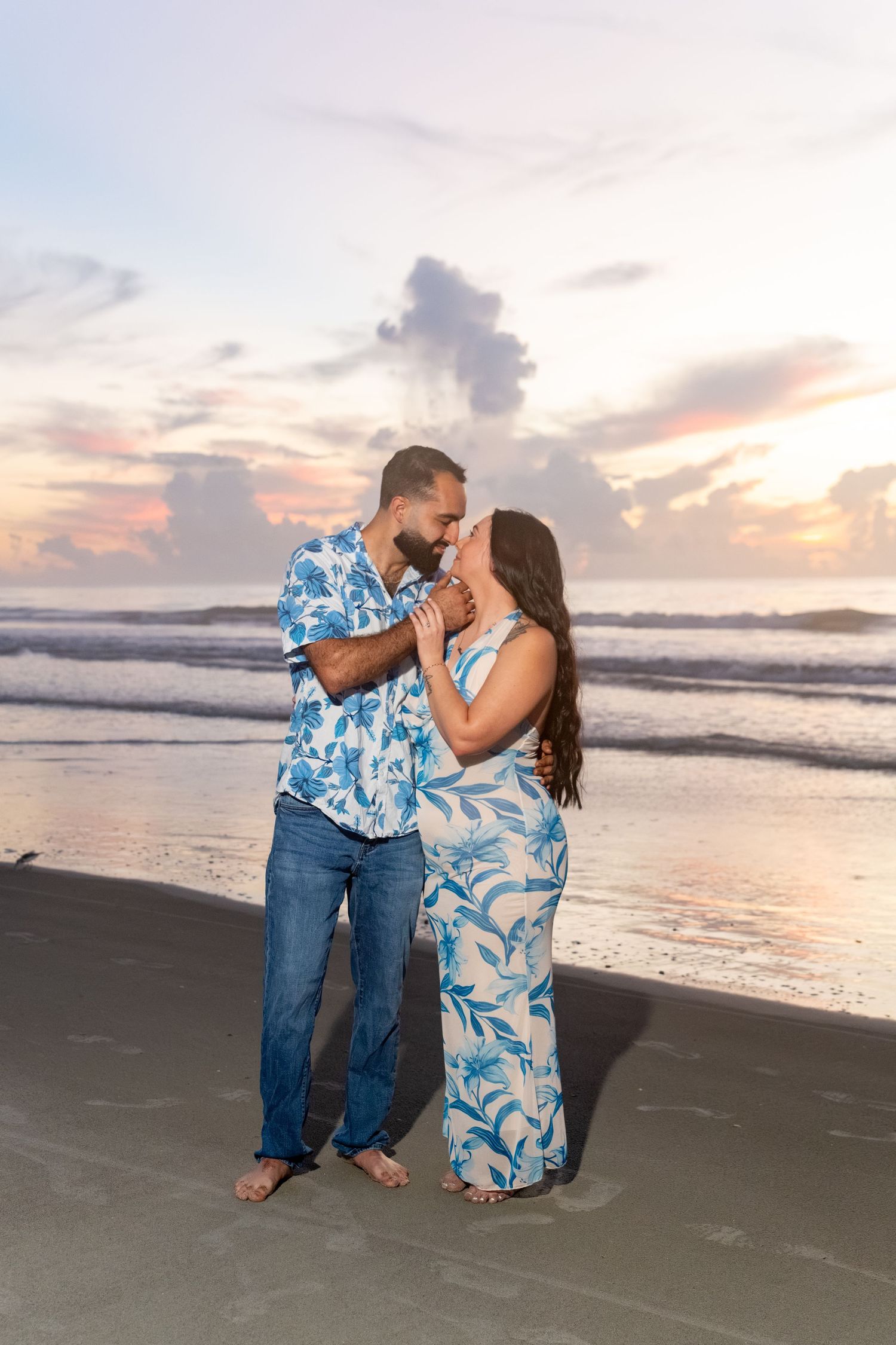 Couple in matching blue floral outfits sharing a romantic moment on the beach at sunset.