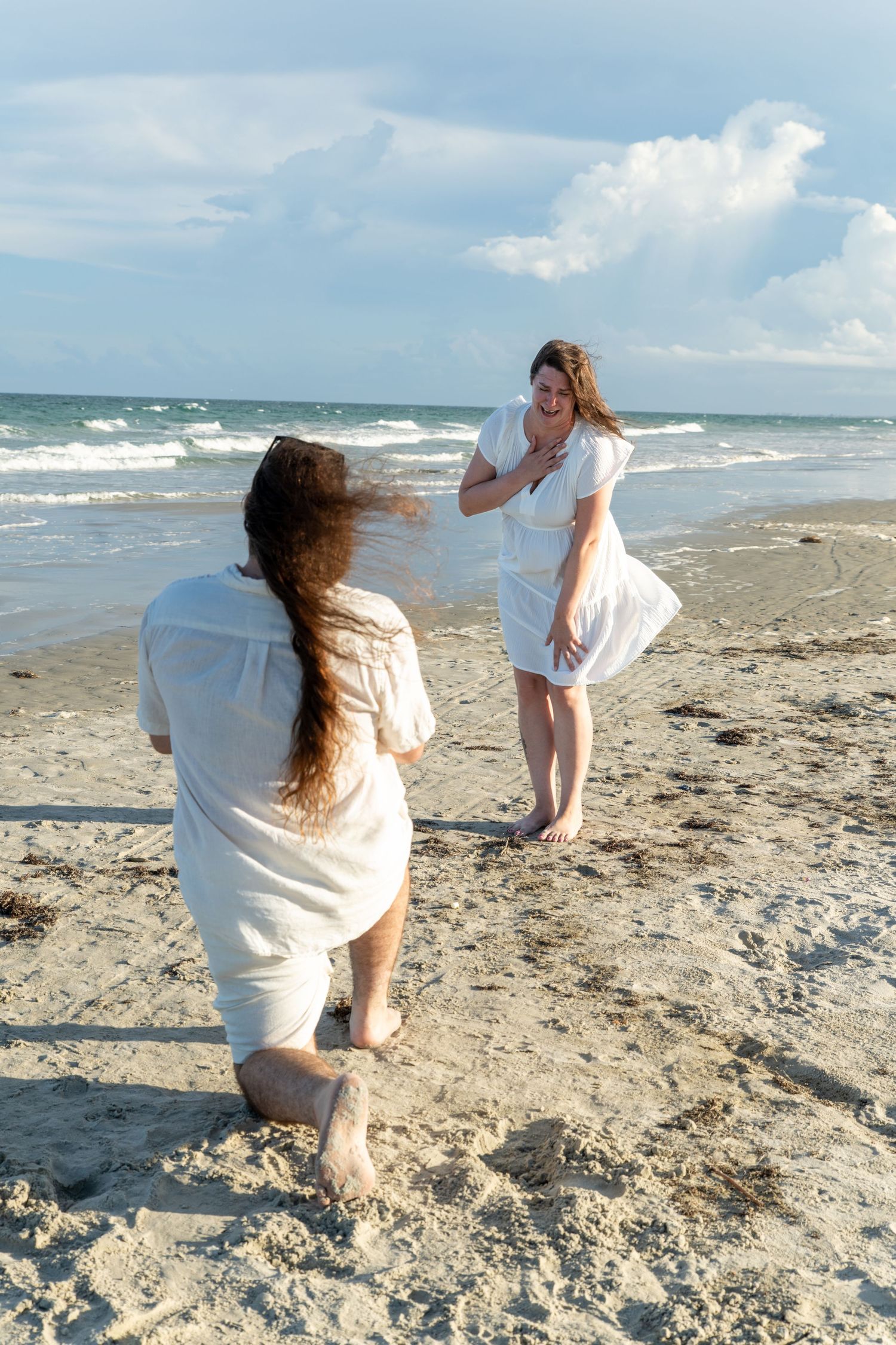 Two women in white dresses enjoy a breezy beach day, posing on sandy shores with ocean waves in the background.