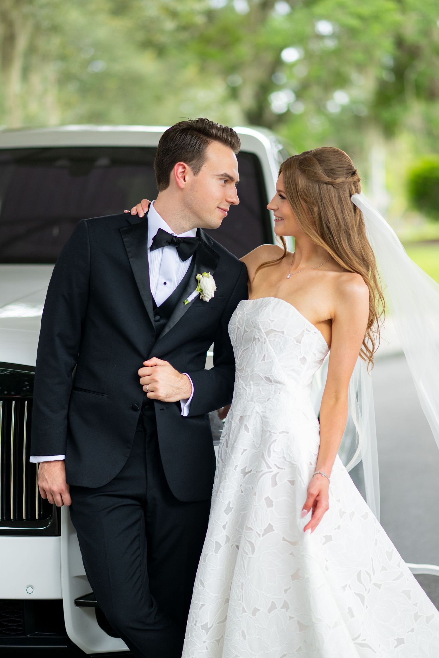 Bride and groom posing together in front of a white luxury car, wearing wedding attire with veil and tuxedo.
