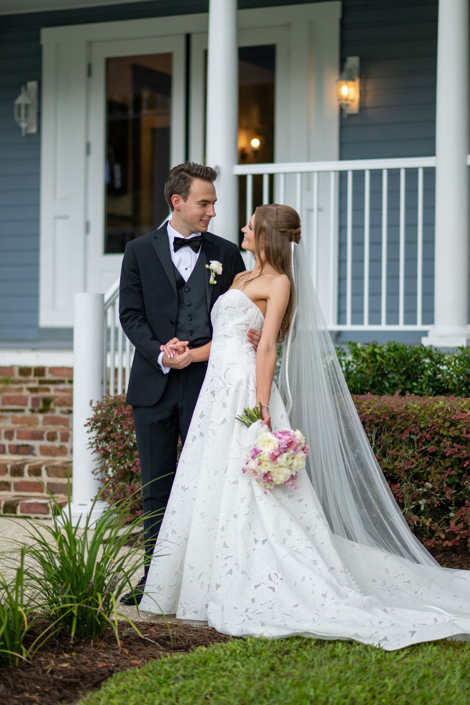 Bride and groom embracing in front of a blue house, she holding a pink bouquet in a strapless lace gown with veil.