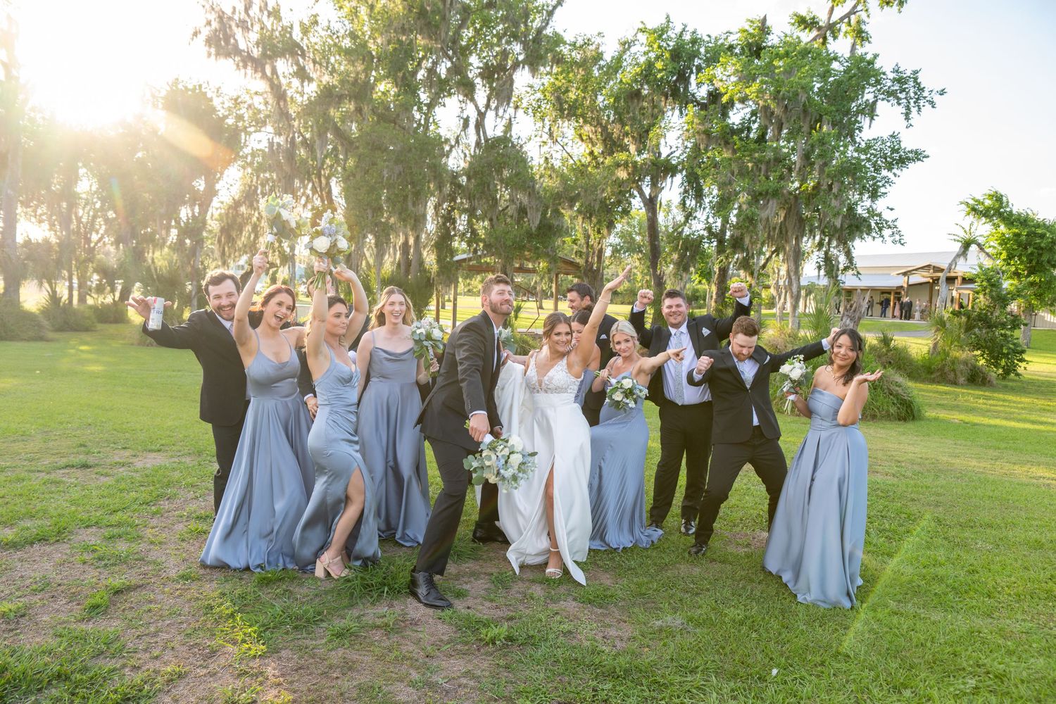 Wedding party in dusty blue dresses and black tuxedos celebrating outdoors under oak trees in golden sunlight.
