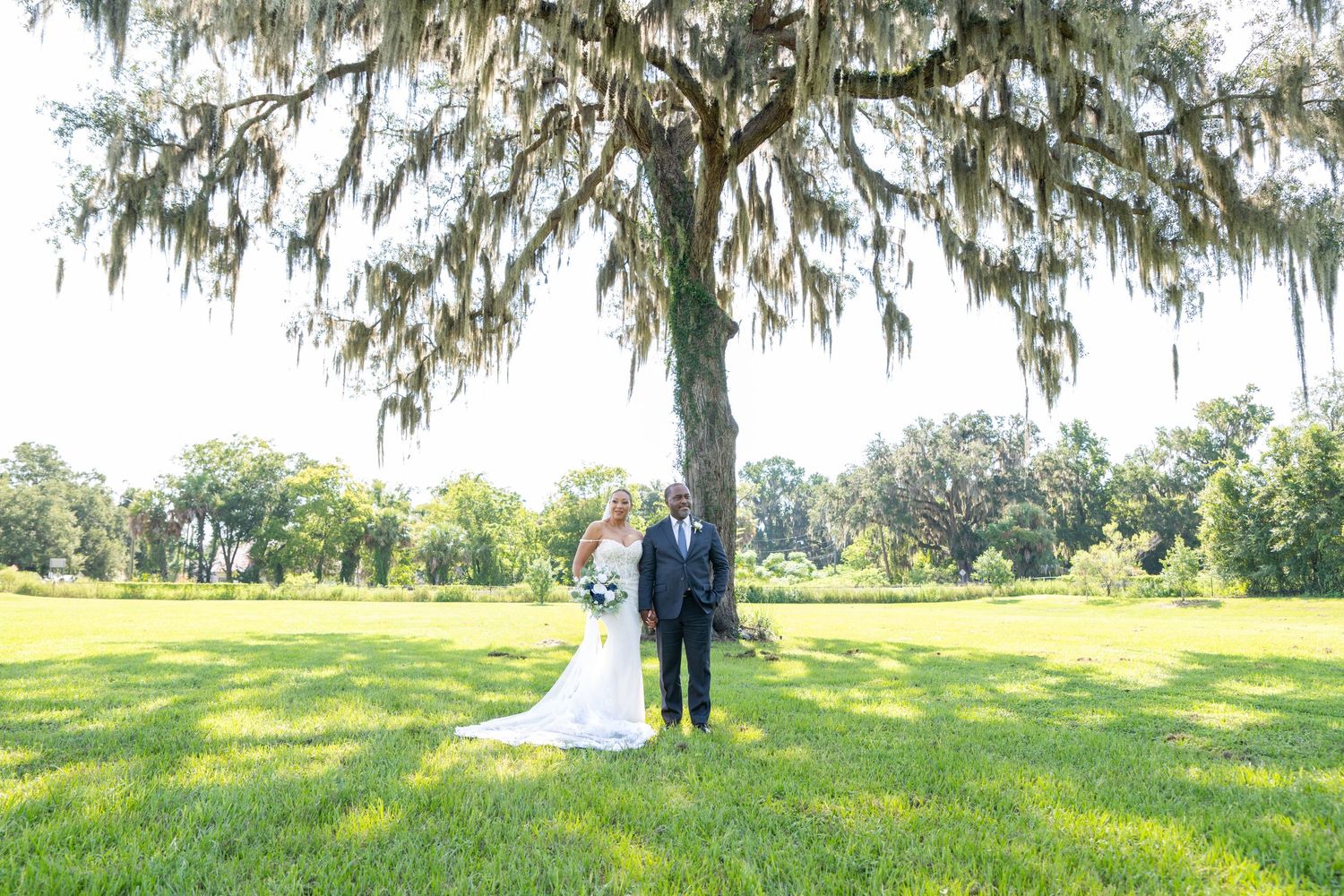 Bride and groom stand beneath a moss-draped oak tree on a lush green lawn during an outdoor wedding ceremony.