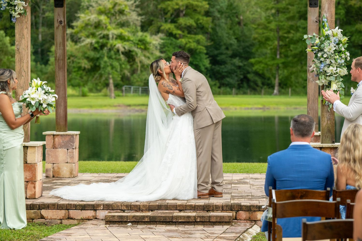 Bride and groom share first kiss at outdoor wedding ceremony by a pond, surrounded by floral arbor and guests.