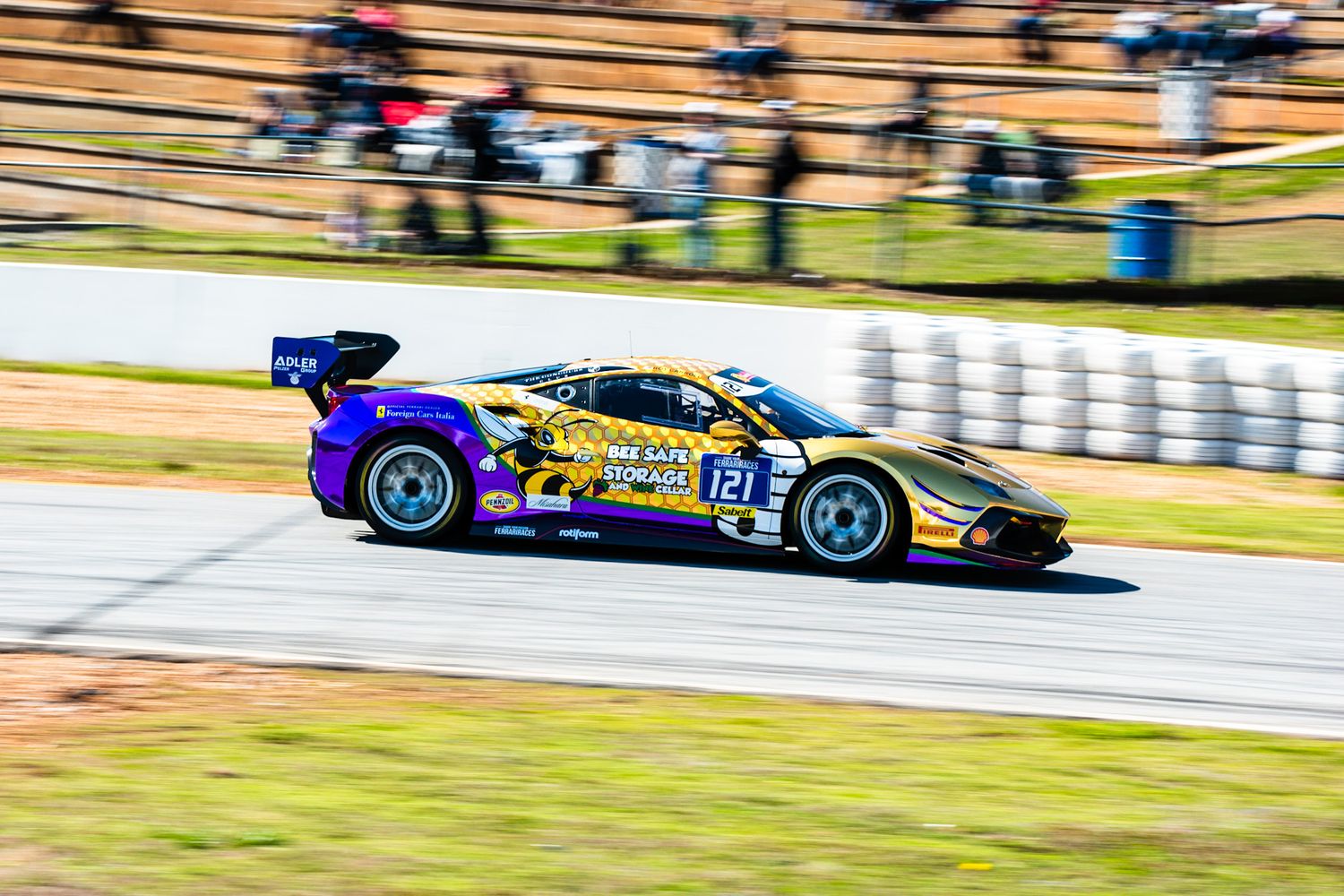 Purple and gold Ferrari 488 GT3 number 121 racing on sunny motorsport circuit.