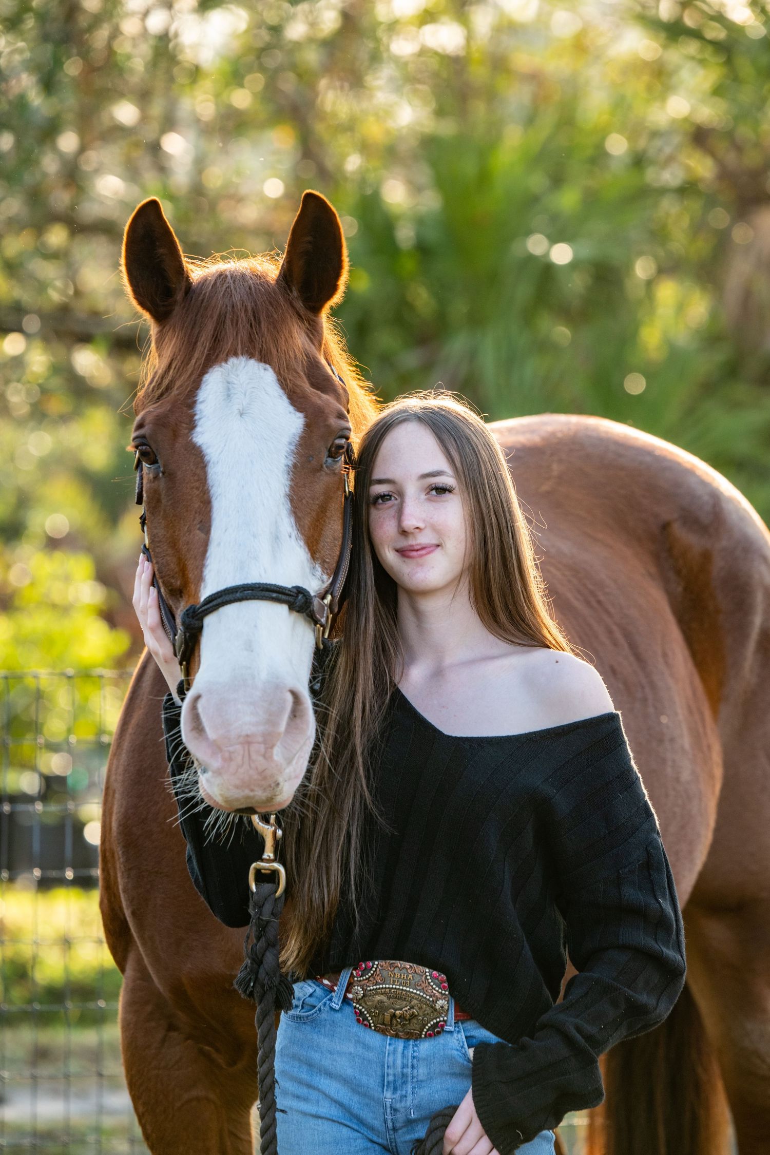 Teen girl with long hair posing beside chestnut horse with white blaze in nature.