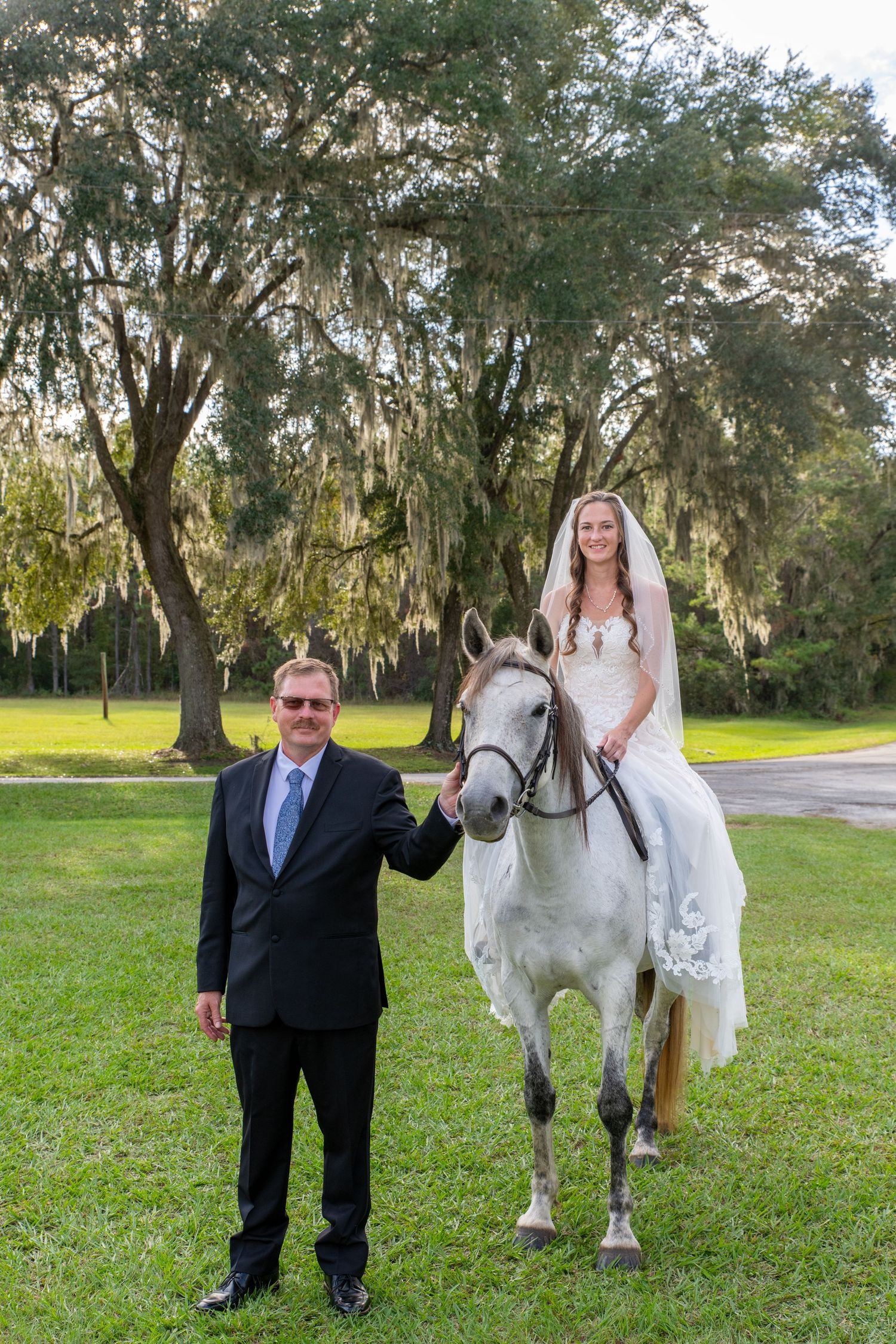 Bride in white wedding gown sits atop a white horse while groom in black suit stands beside her on green lawn.