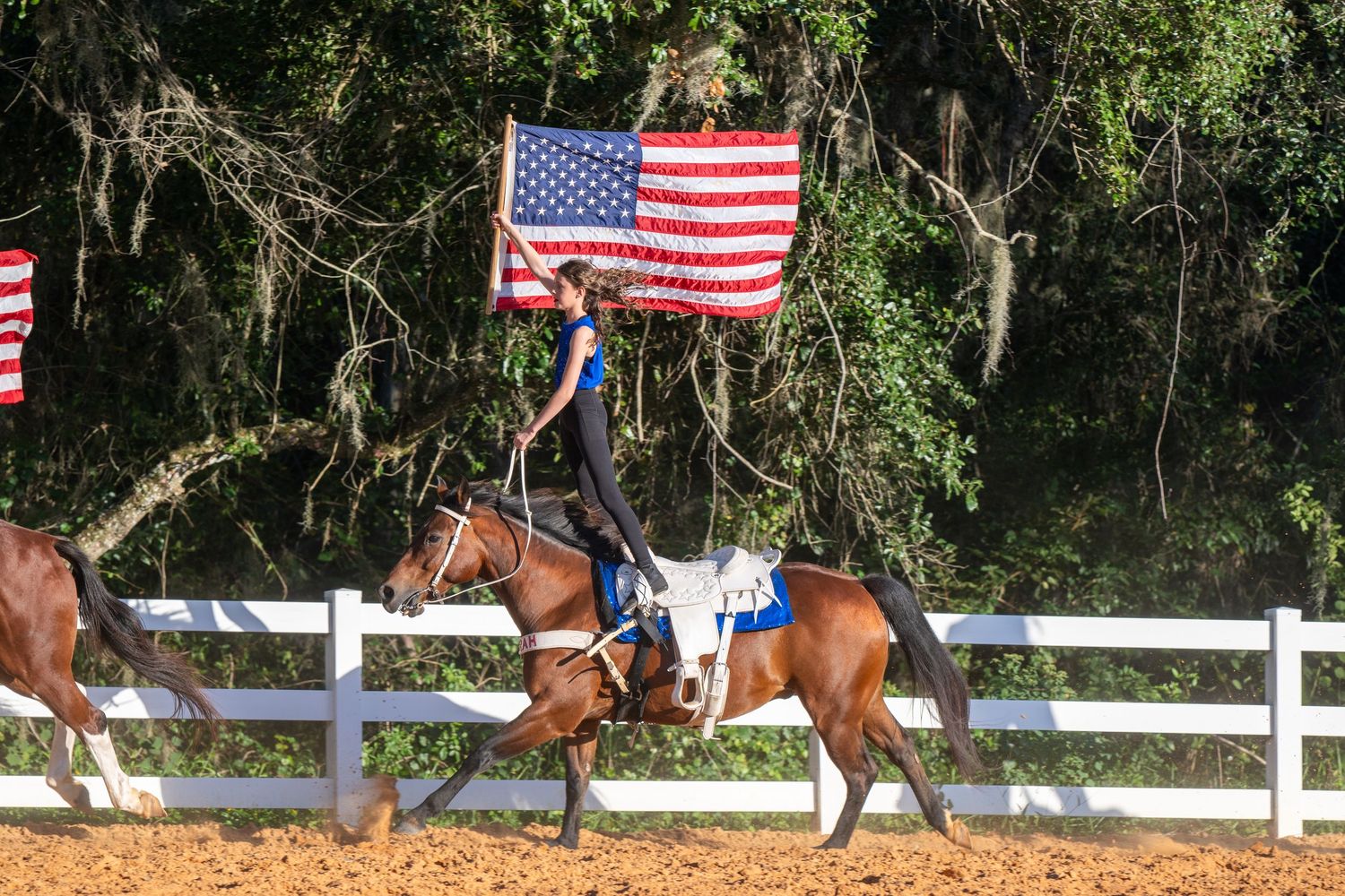 A rider on horseback carries an American flag in an outdoor equestrian arena lined with white fencing.